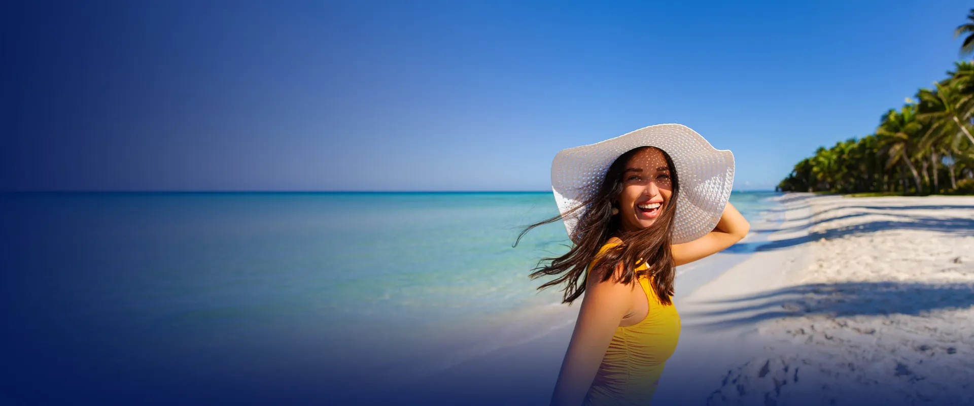 Smiling woman in a yellow swimsuit and white sun hat on a tropical beach with palm trees and turquoise water under a clear blue sky.
