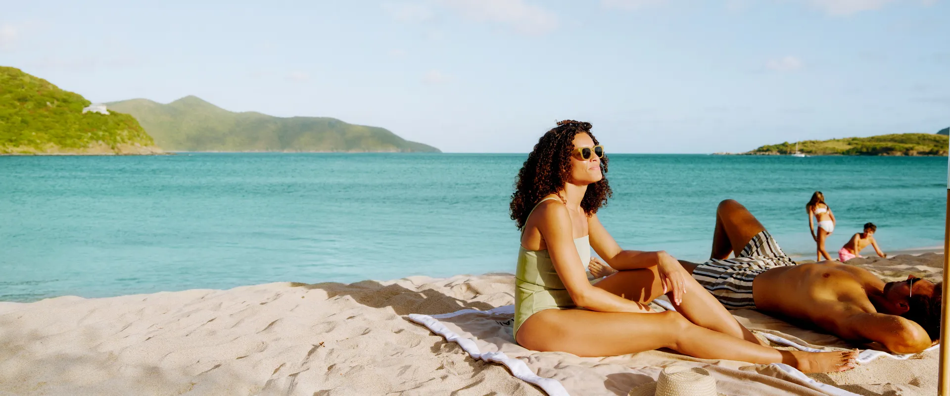 A woman in a swimsuit and sunglasses relaxes on a sandy beach, with turquoise water and green hills in the background.