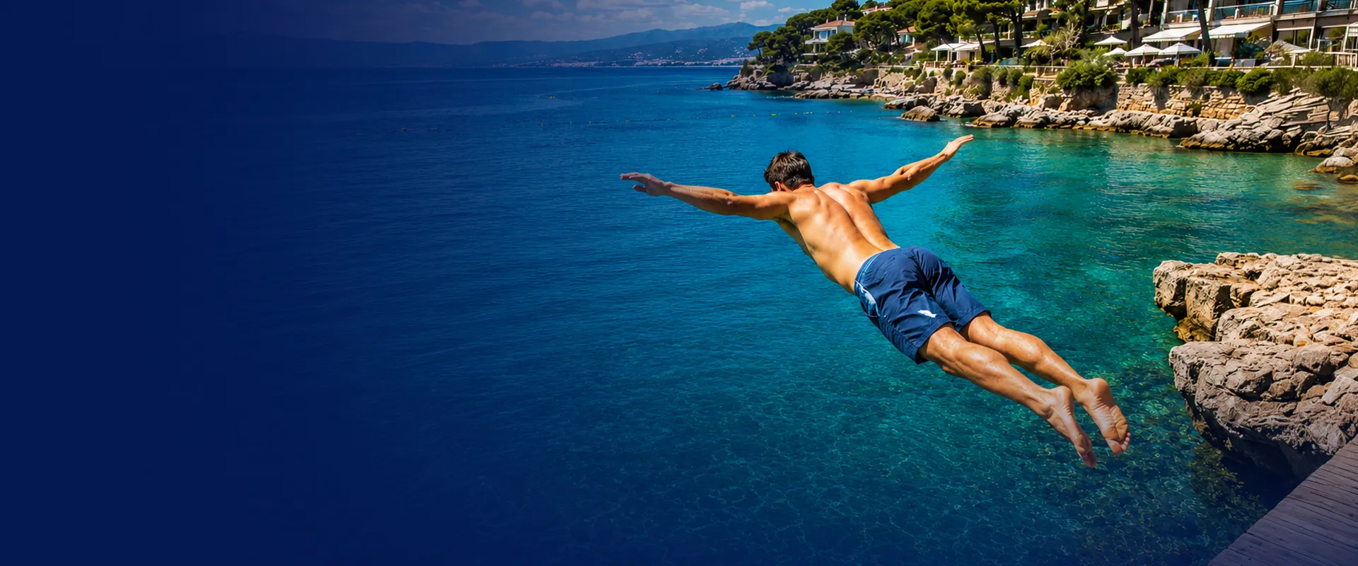 Man diving off a rocky shoreline into clear blue water, with a coastal landscape and houses in the background.