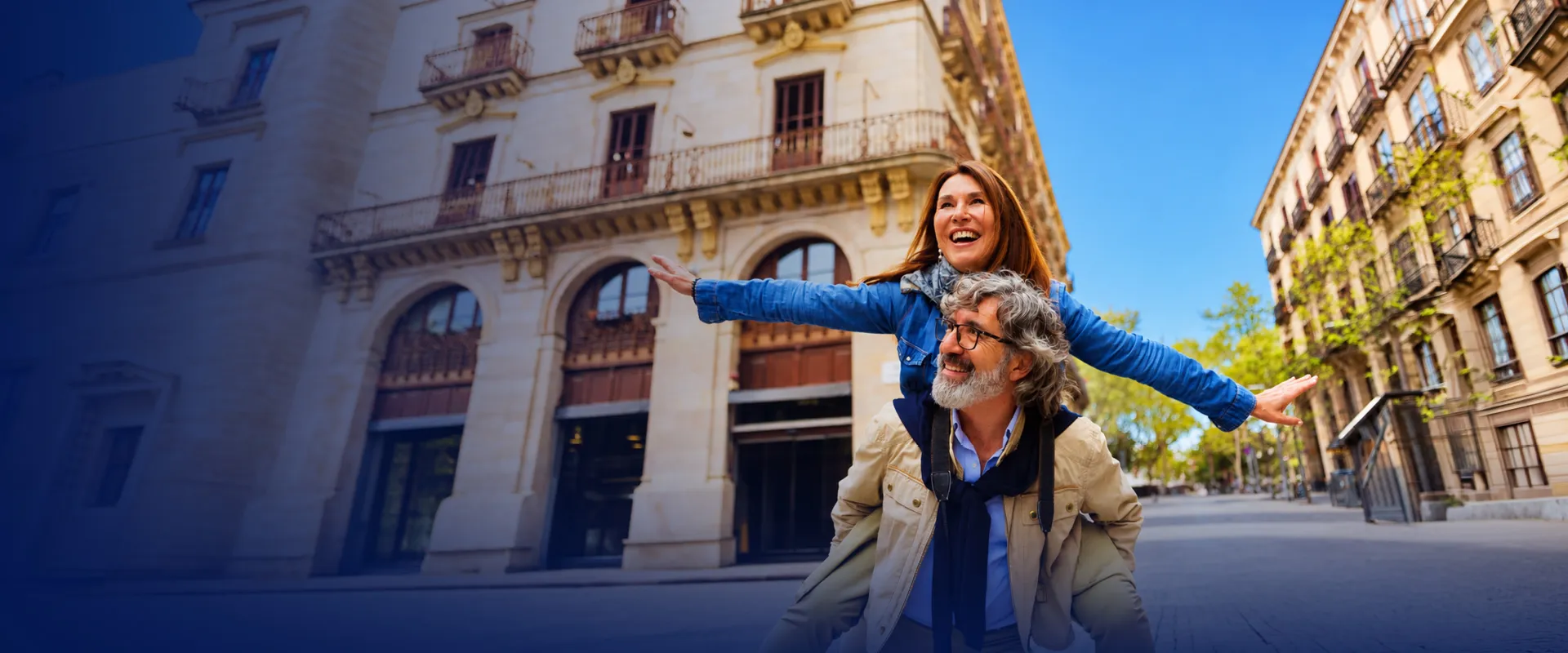 A woman joyfully rides on a man's back as they walk down a bright, picturesque street with elegant buildings under a clear blue sky.