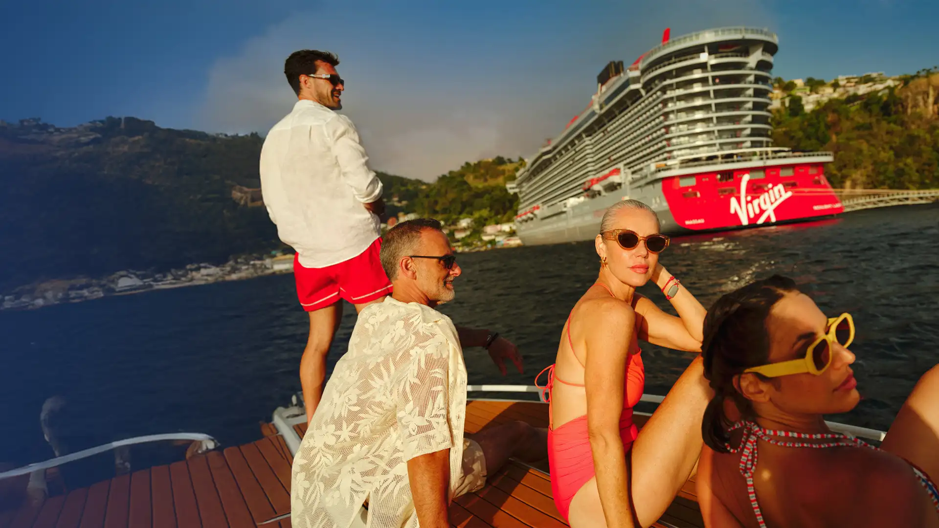 People on a boat relaxing and looking towards a large red Virgin cruise ship near a scenic coastline.