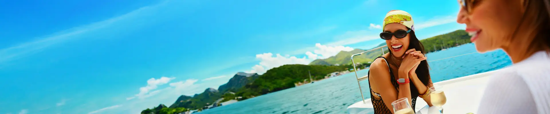 Two women enjoying drinks on a boat, with clear blue skies, mountains, and ocean in the background.