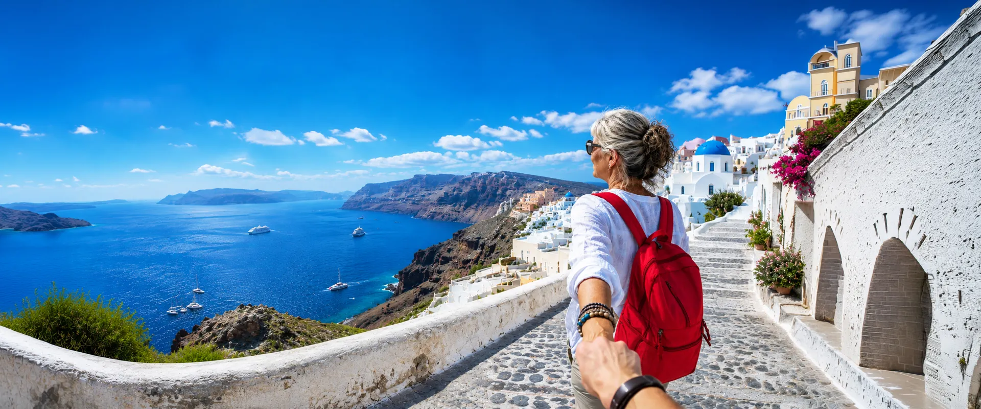 Woman with red backpack, holding hand, walks along cobblestone path overlooking sea and white buildings, Santorini, Greece.