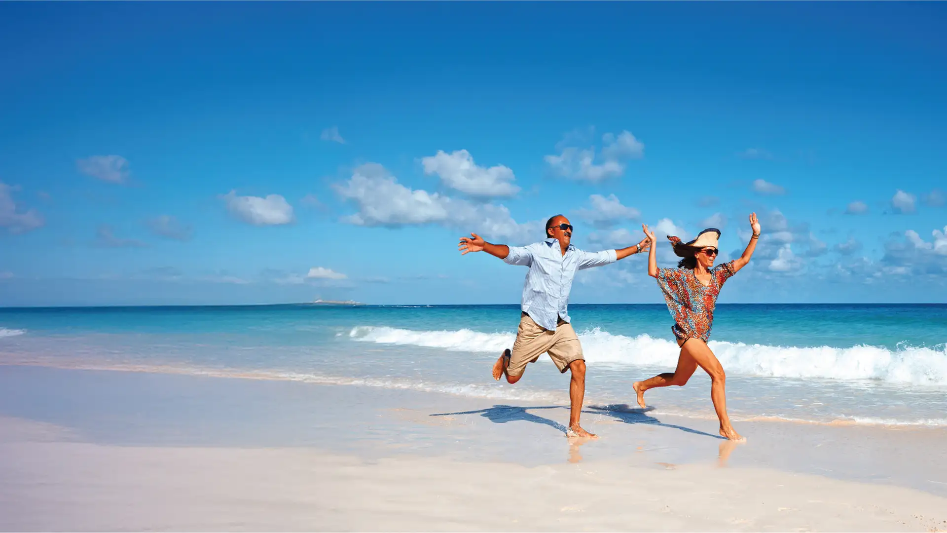 A joyful couple runs along a sunny, sandy beach with clear blue skies and ocean waves in the background.