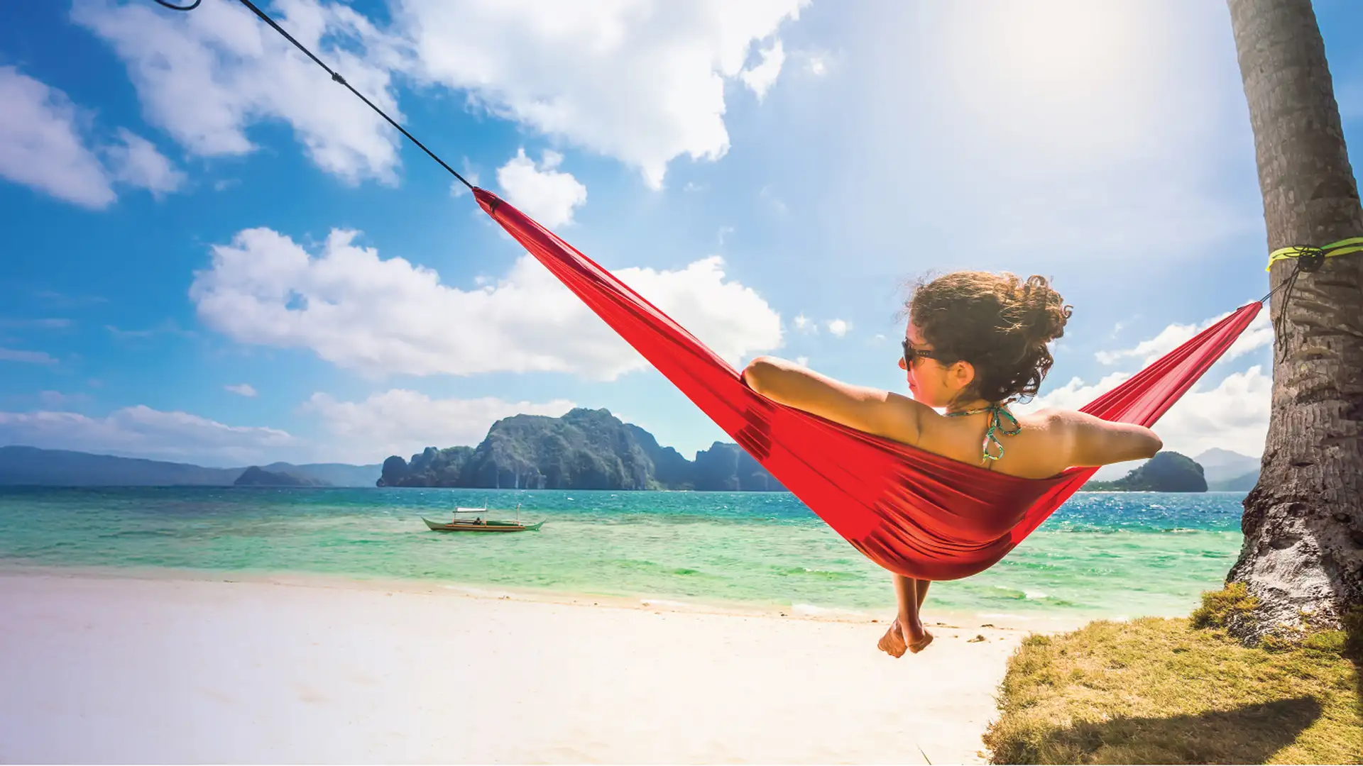 A woman relaxes in a red hammock tied to a palm tree on a sunny beach, overlooking turquoise waters and distant islands.