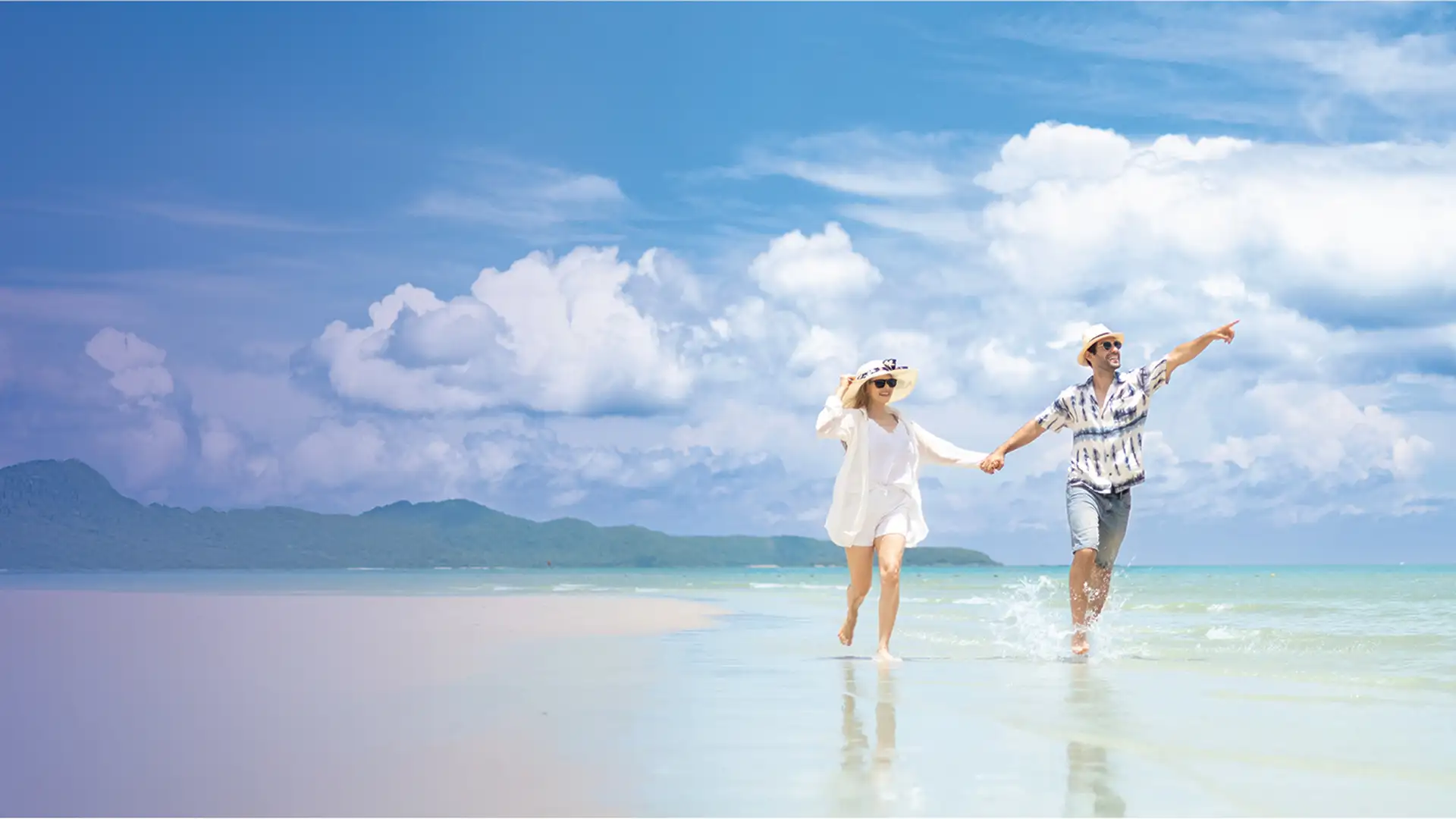 A couple in summer attire joyfully walks hand in hand along a serene beach, with clear blue skies and distant mountains in the background.