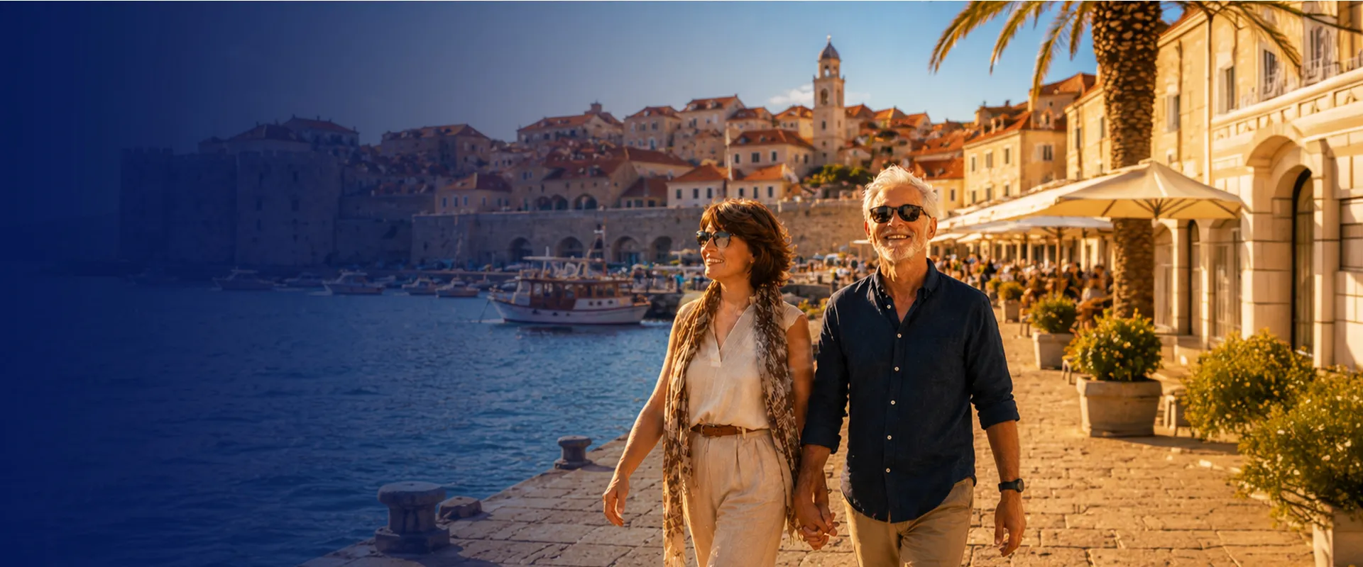 A couple holds hands, smiling while walking by the waterfront in a sunlit, historical European town with boats in the harbor.