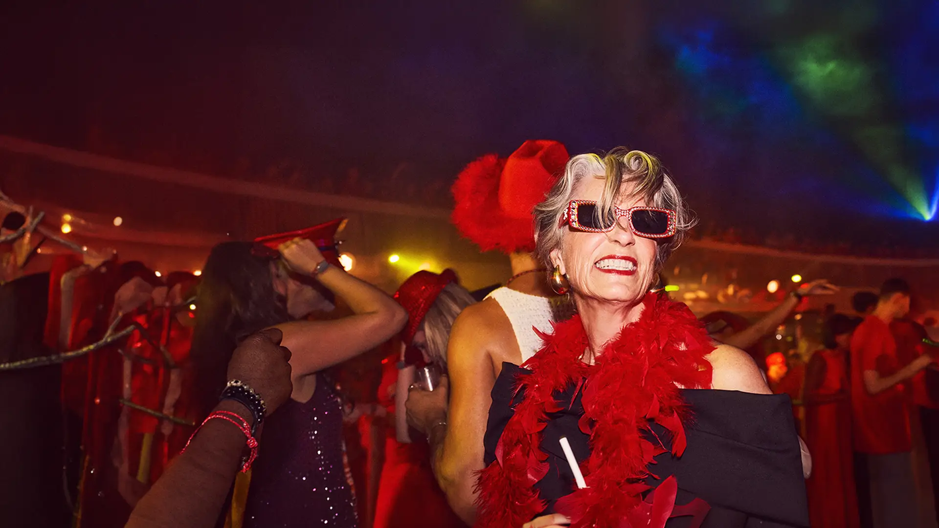 Elderly woman wearing party attire, red feather boa, and sunglasses, dancing joyfully at a lively, colorful dance party.