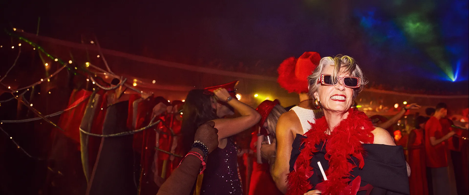 Elderly woman in red boa and funky glasses dances energetically at a lively, festive party with colorful lights and decorations.