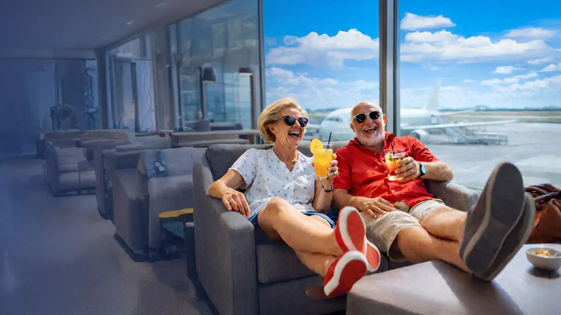 Elderly couple relaxing in an airport lounge with drinks and snacks, sitting on a sofa, enjoying a view of the runway and blue sky outside.