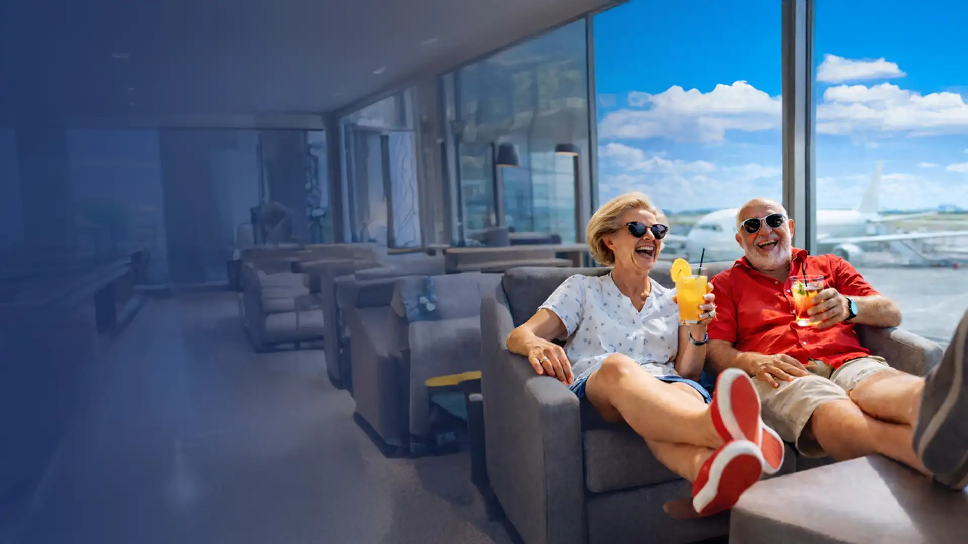 Elderly couple in casual clothes relax in an airport lounge with drinks, smiling, and looking out at the runway and blue sky.