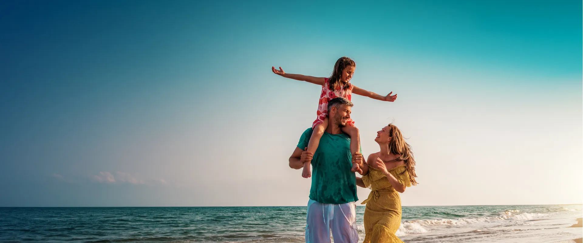 A family enjoys a sunny day on the beach; a man carries a child on his shoulders while a woman smiles beside them. The ocean is in the background.