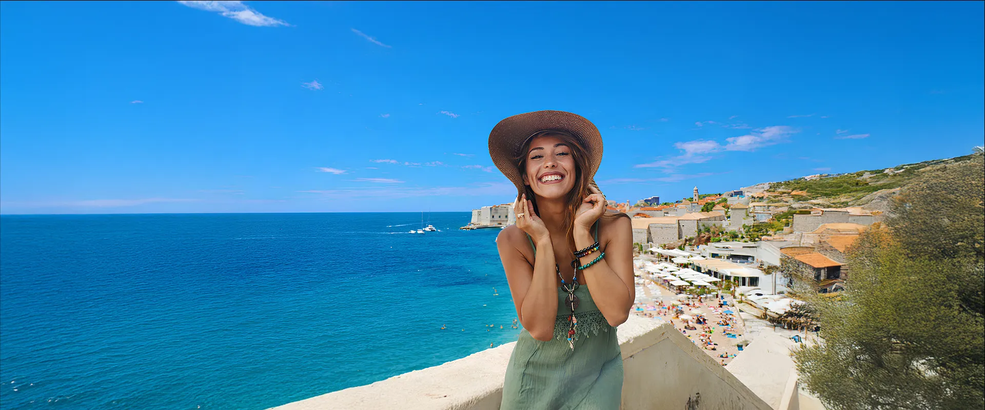 Smiling woman in a sunhat poses on a coastal balcony, with a vibrant blue ocean and a crowded beach in the background under a clear sky.