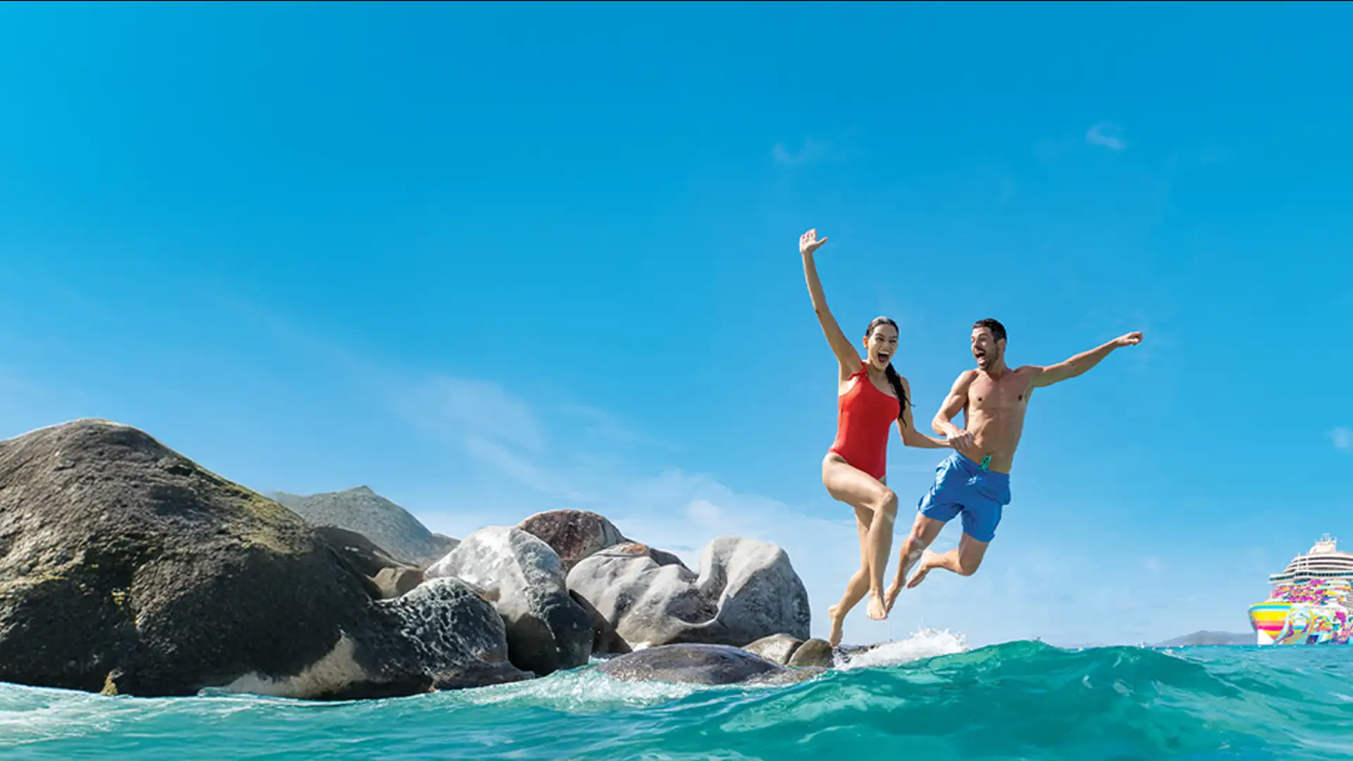 A man and woman joyfully jump from rocks into the ocean, with a colorful cruise ship in the background under a clear blue sky.