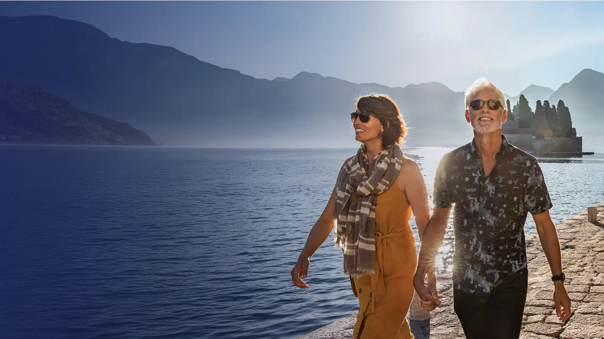 A couple holding hands, wearing sunglasses, walks on a stone path beside a tranquil lake, with mountains in the background under a clear blue sky.