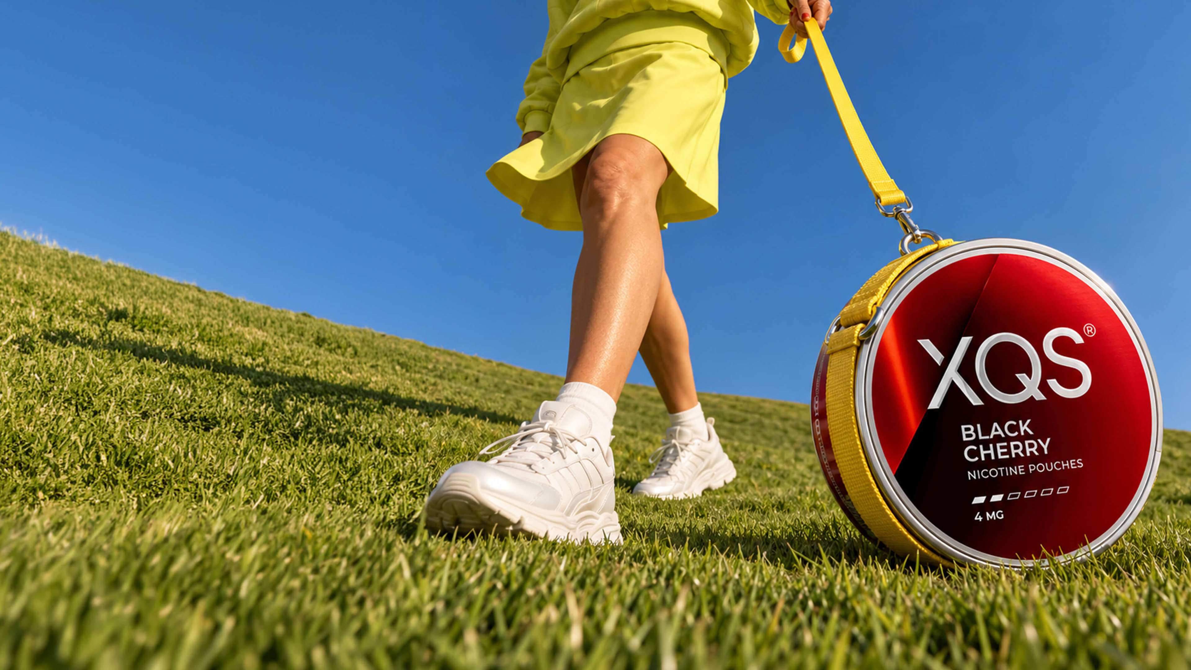 Person in yellow outfit and white sneakers walking on grassy hill, carrying large XQS Black Cherry nicotine pouches container with yellow strap.