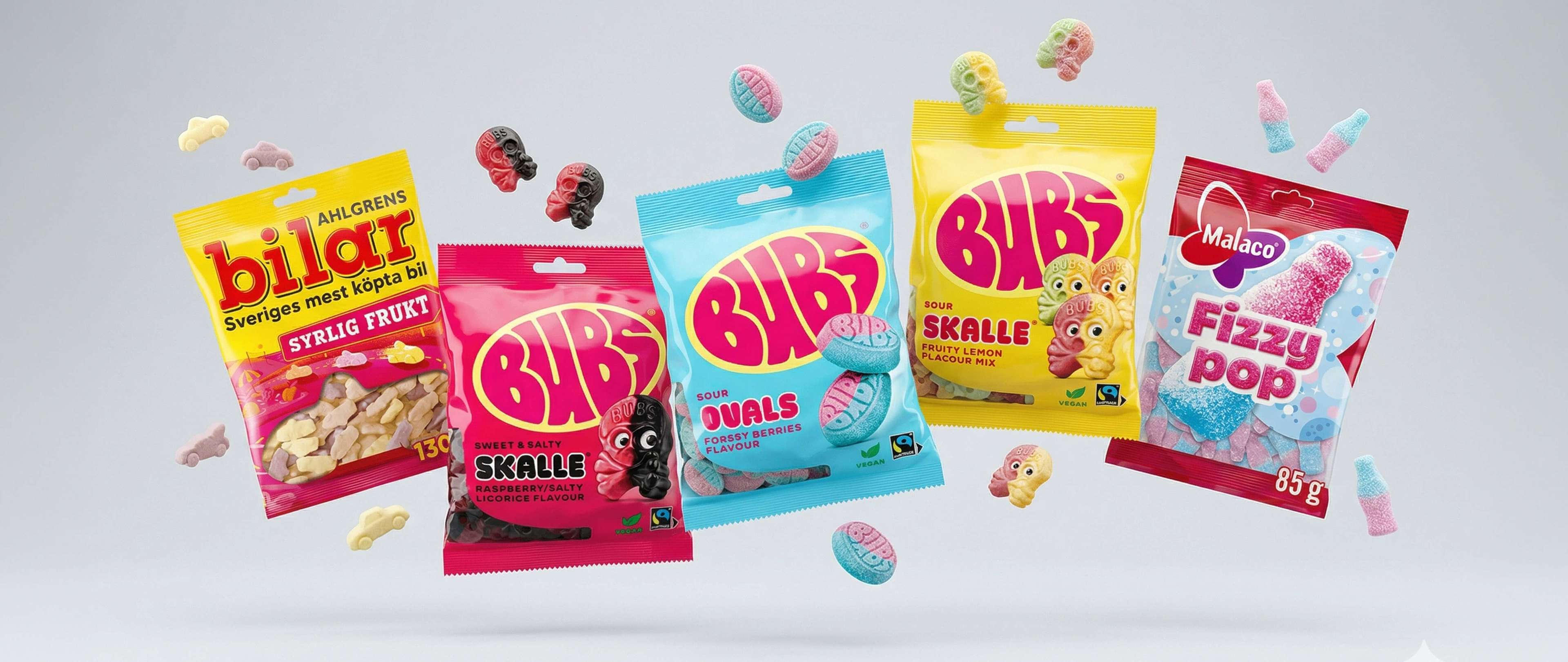 Assorted colorful swedish candy packages and scattered sweets, including Bubs Sour Skalle, Bilar, and Malaco Fizzy Pop, against a light background.