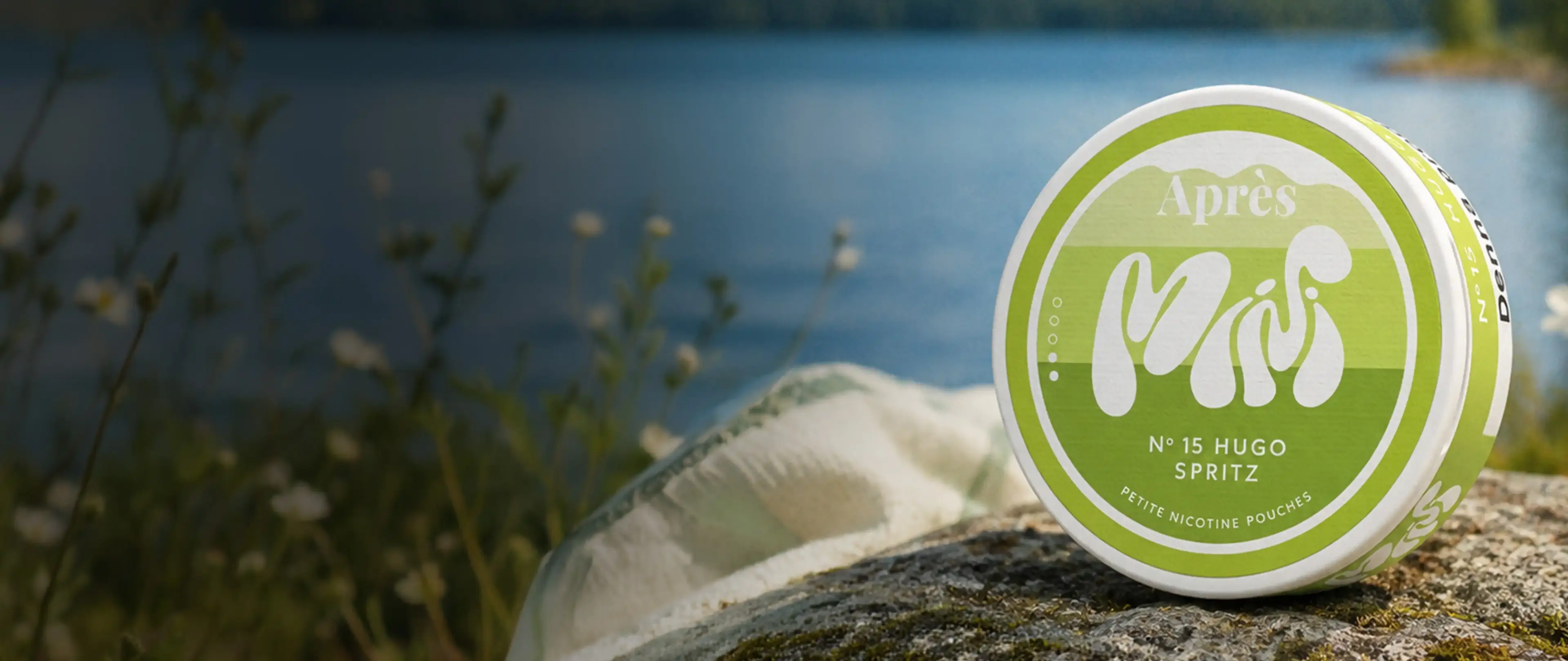 Container of Après Hugo Spritz nicotine pouches on a rock by a lake, with blurred wildflowers in the background.