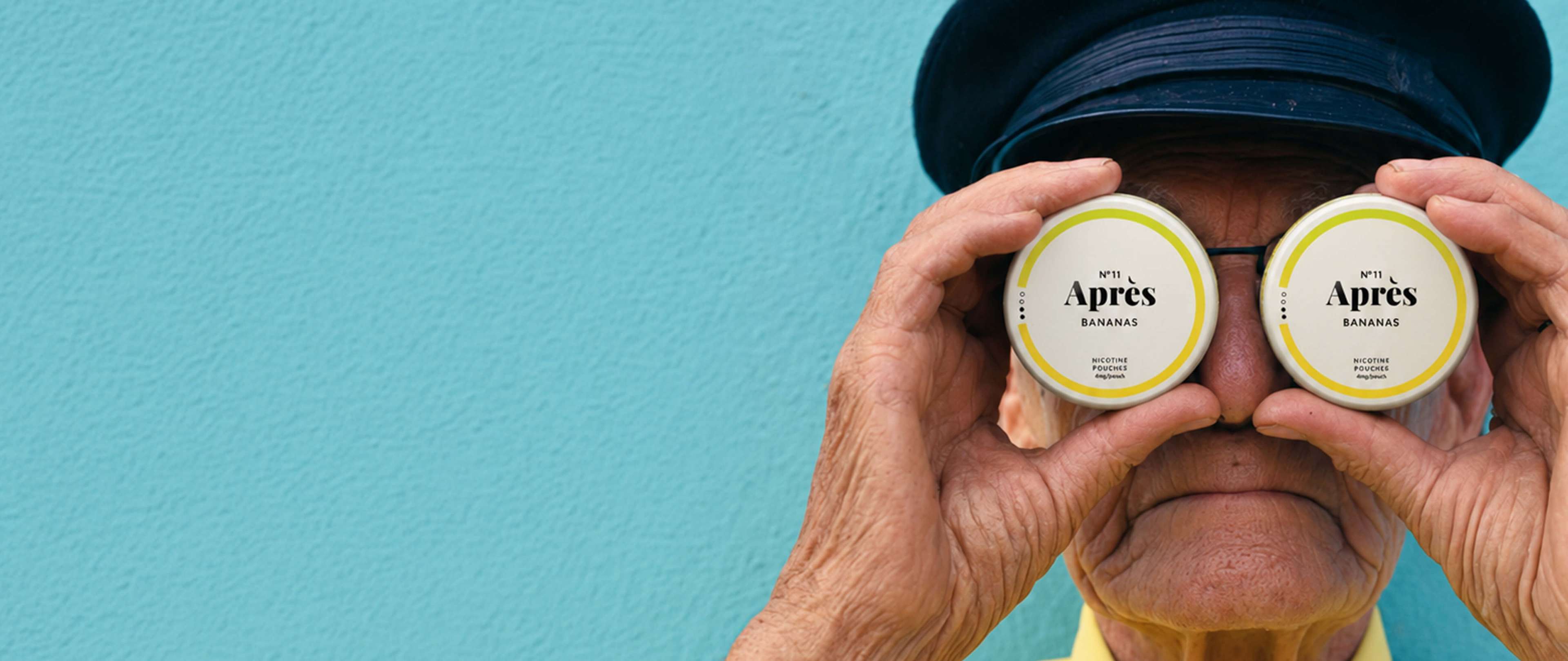 Person wearing a cap holds two cans labeled "Après Bananas Nicotine Pouches" over their eyes against a blue background.