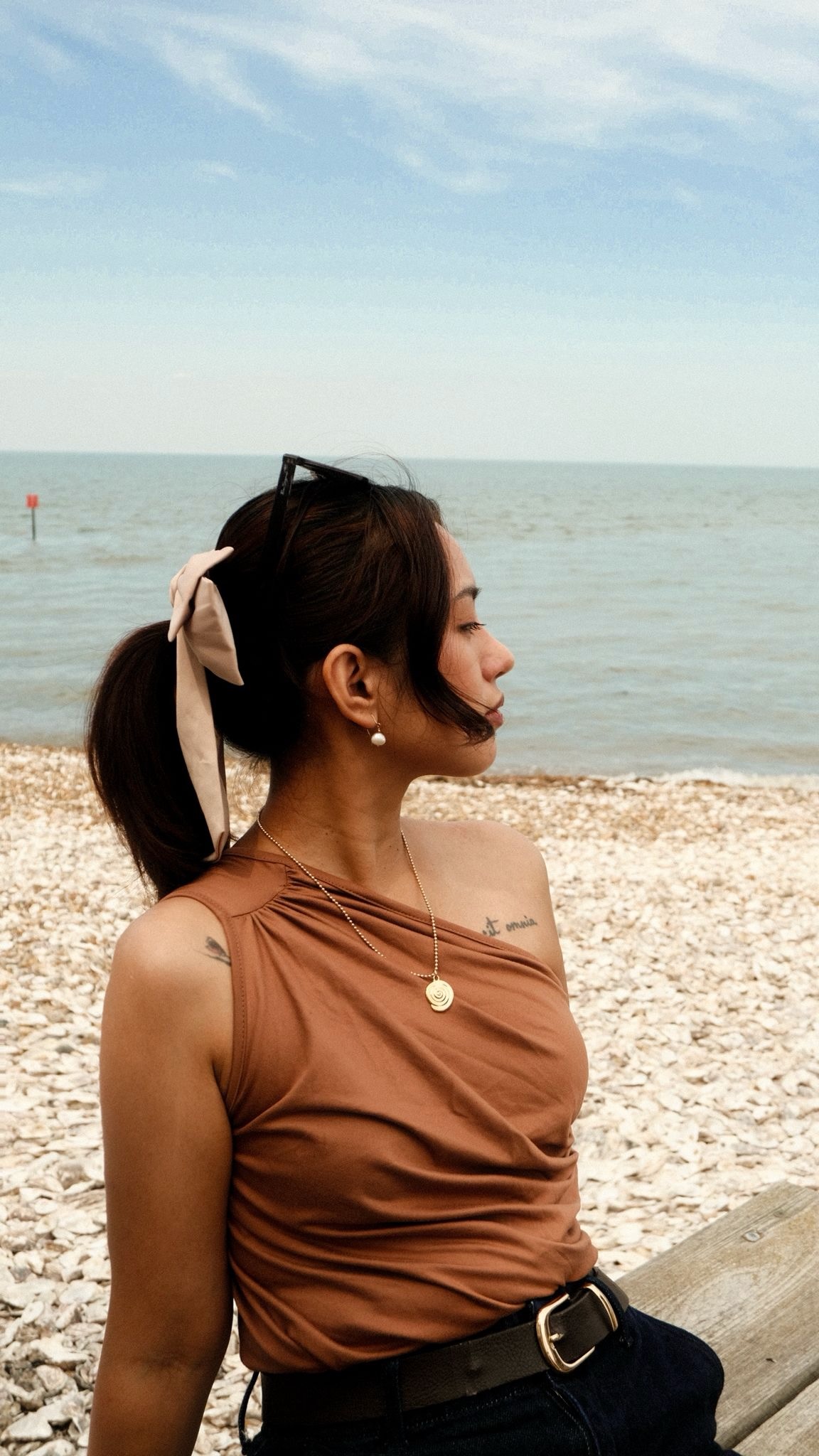 Woman with sunglasses on head and ribbon in hair, wearing a brown top, stands by a pebbly beach, gazing at the sea under a blue sky.