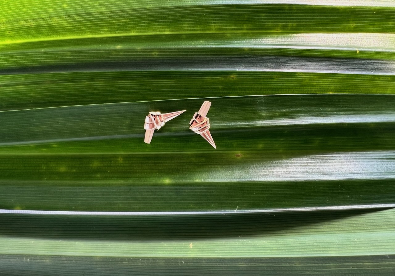 Two small, intricately designed pandan knot gold earrings on a background of layered green leaves.