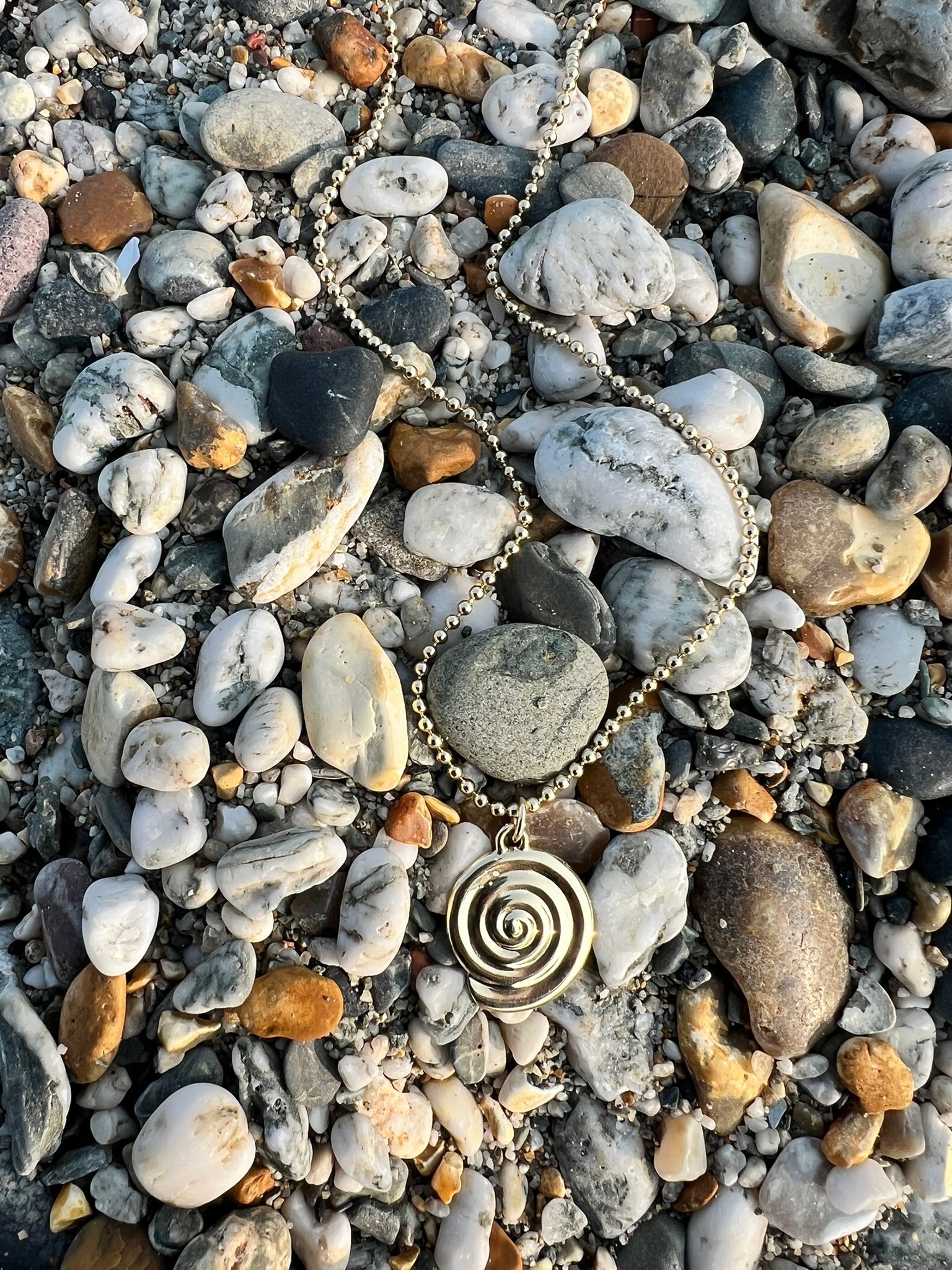 A gold spiral pendant on a beaded chain lies on a bed of multicolored pebbles and stones.