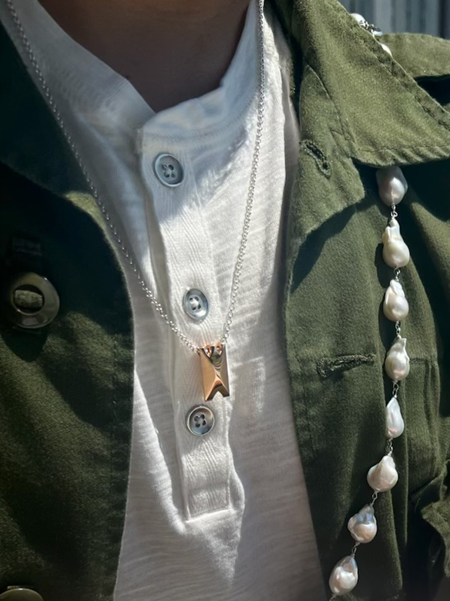 Person wearing a white buttoned shirt, green jacket with pearl details, and a silver necklace with a gold pendant.