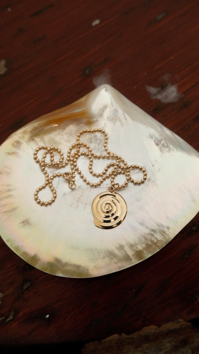 Gold spiral pendant necklace with a beaded chain, resting on an iridescent shell on a wooden surface.