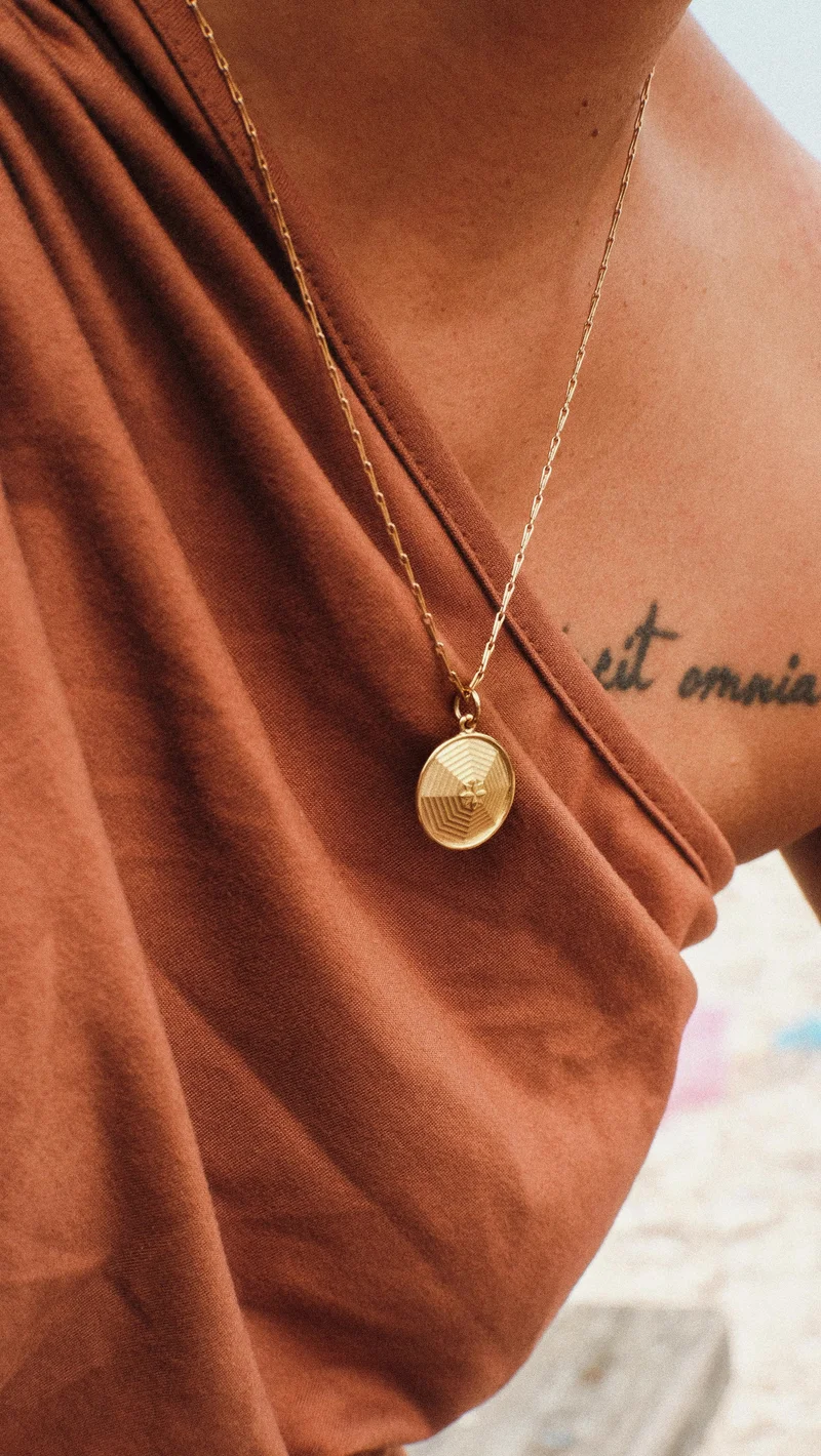 A person wearing a rust-colored top and a gold pendant necklace. A partial tattoo is visible on their shoulder.