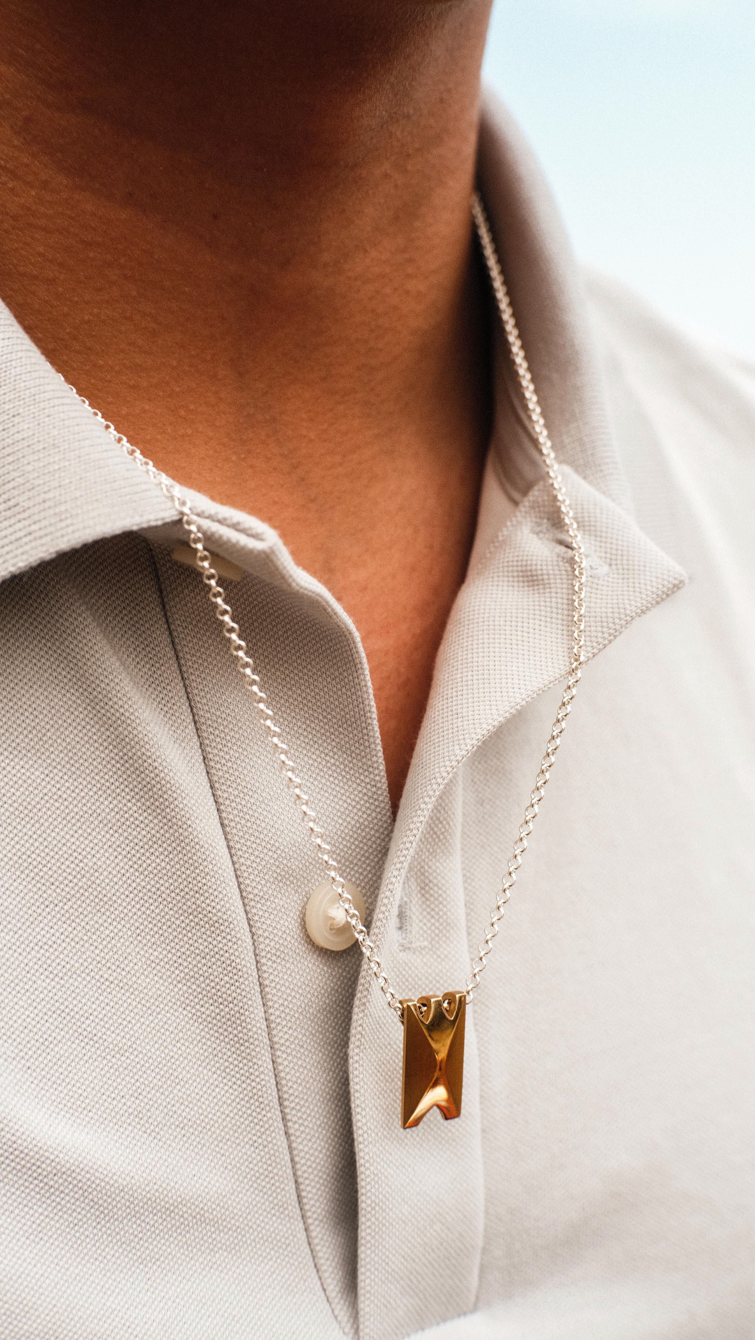 A person wearing a light gray collared shirt and a silver chain necklace with a gold rectangular pendant.
