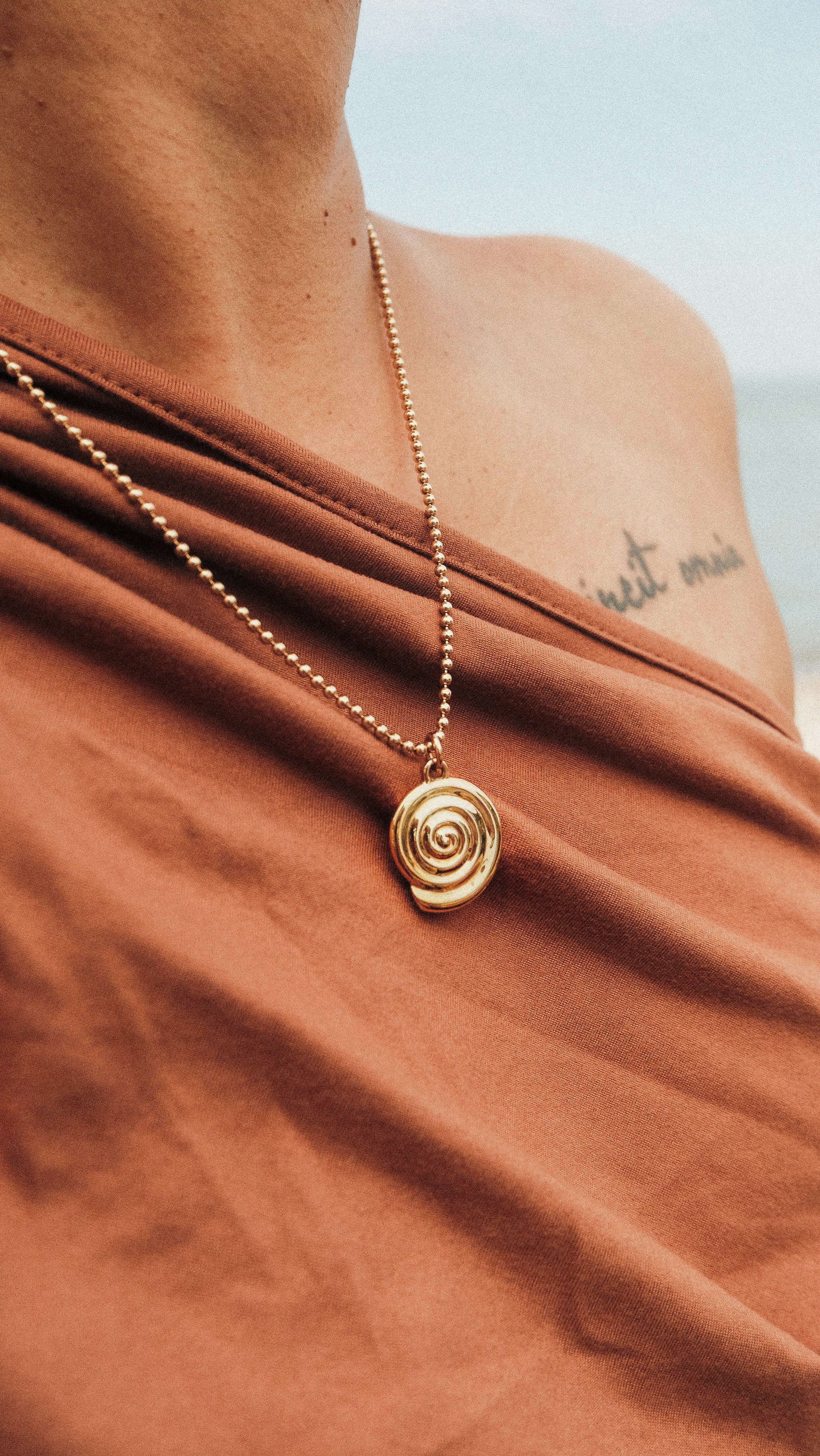 Person wearing a gold spiral pendant necklace over a brown top, with a partial tattoo visible on the shoulder.