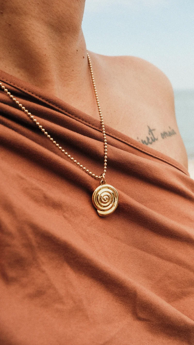 Person wearing a gold spiral pendant necklace over a brown top, with a partial tattoo visible on the shoulder.