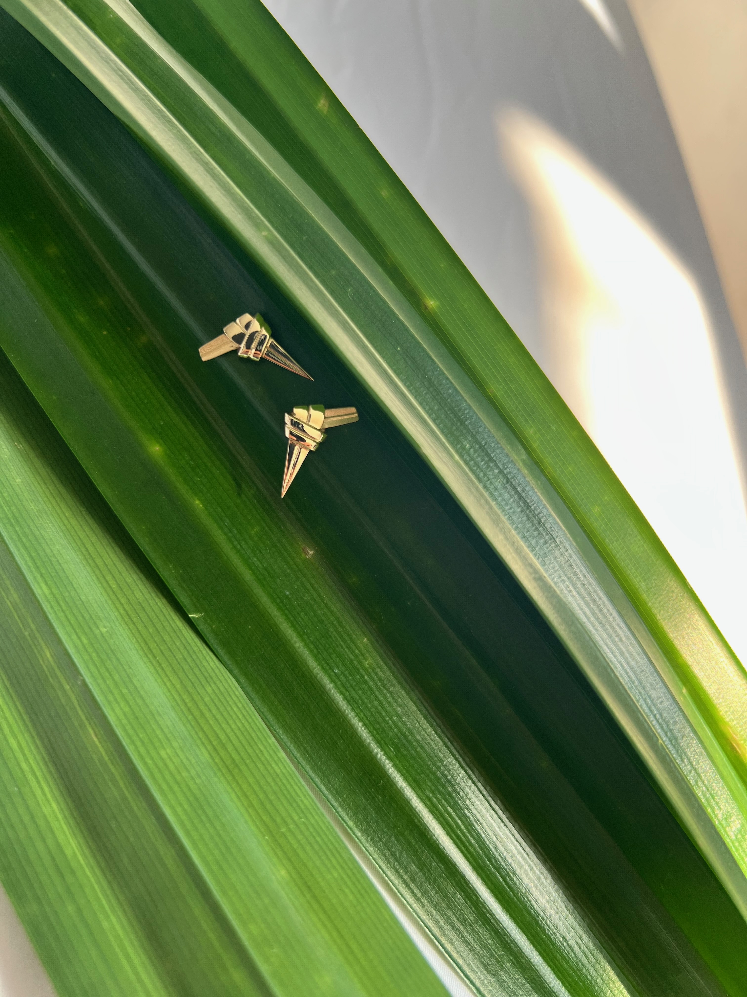 Two gold pandan knot earrings rest on broad, glossy green leaves, with sunlight casting soft shadows.
