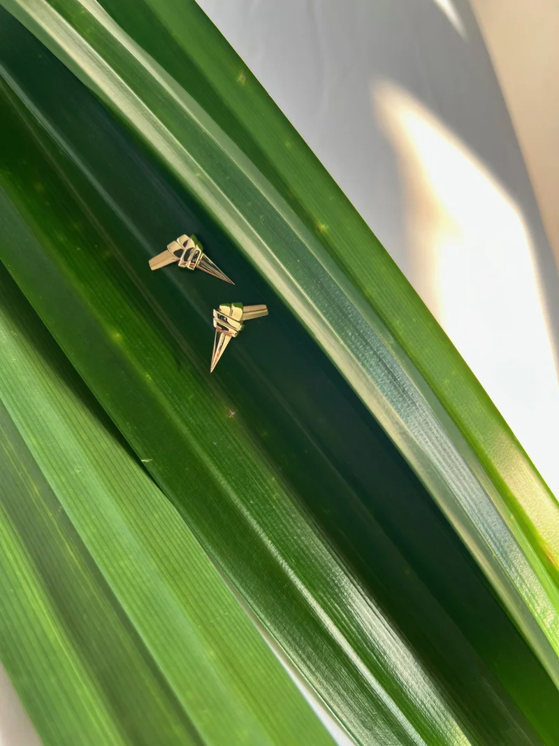 Two gold pandan knot earrings rest on broad, glossy green leaves, with sunlight casting soft shadows.