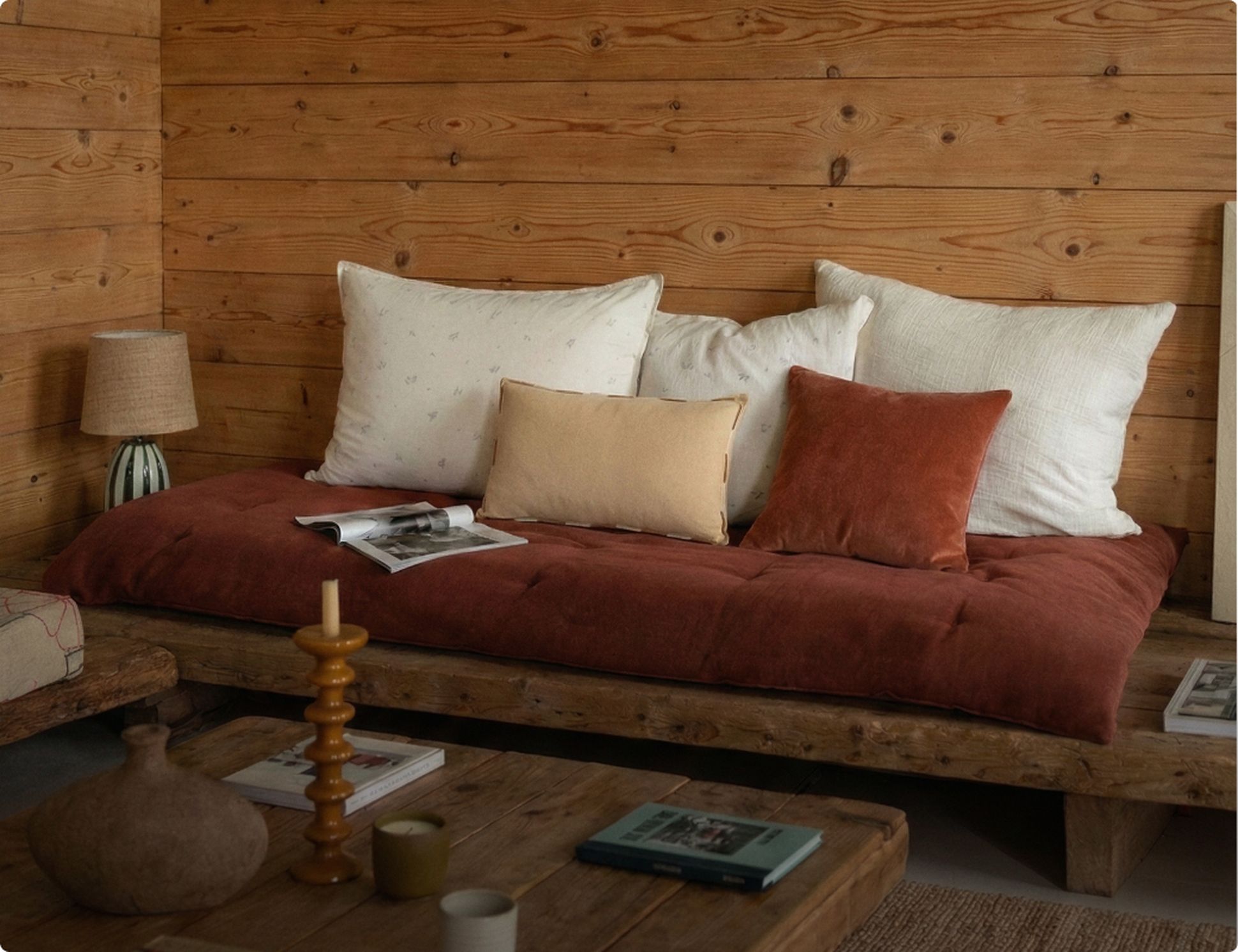 Pièce confortable en bois avec un canapé, des coussins assortis et une table basse rustique avec des livres, une bougie et une lampe pour créer un cadre chaleureux et accueillant.