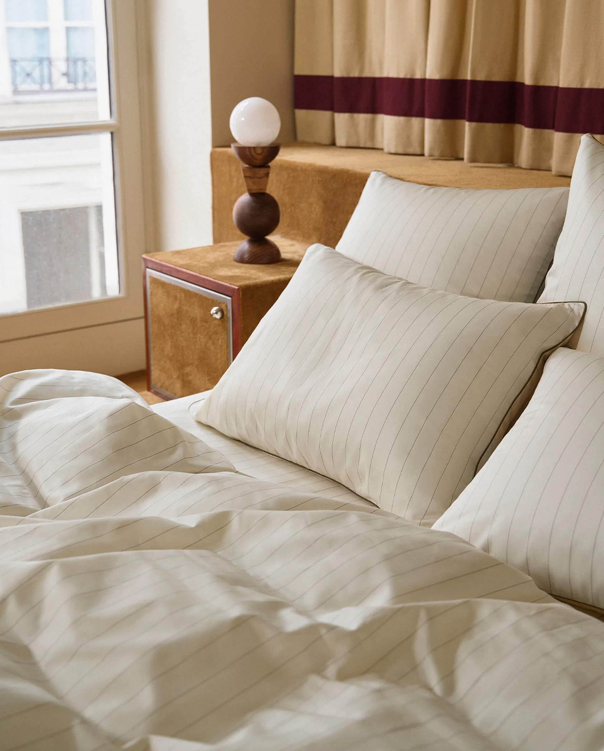 Un lit bien fait, avec des draps et des oreillers rayés de blanc, à côté d'une table de chevet en bois sur laquelle repose une lampe décorative. Une fenêtre en arrière-plan.