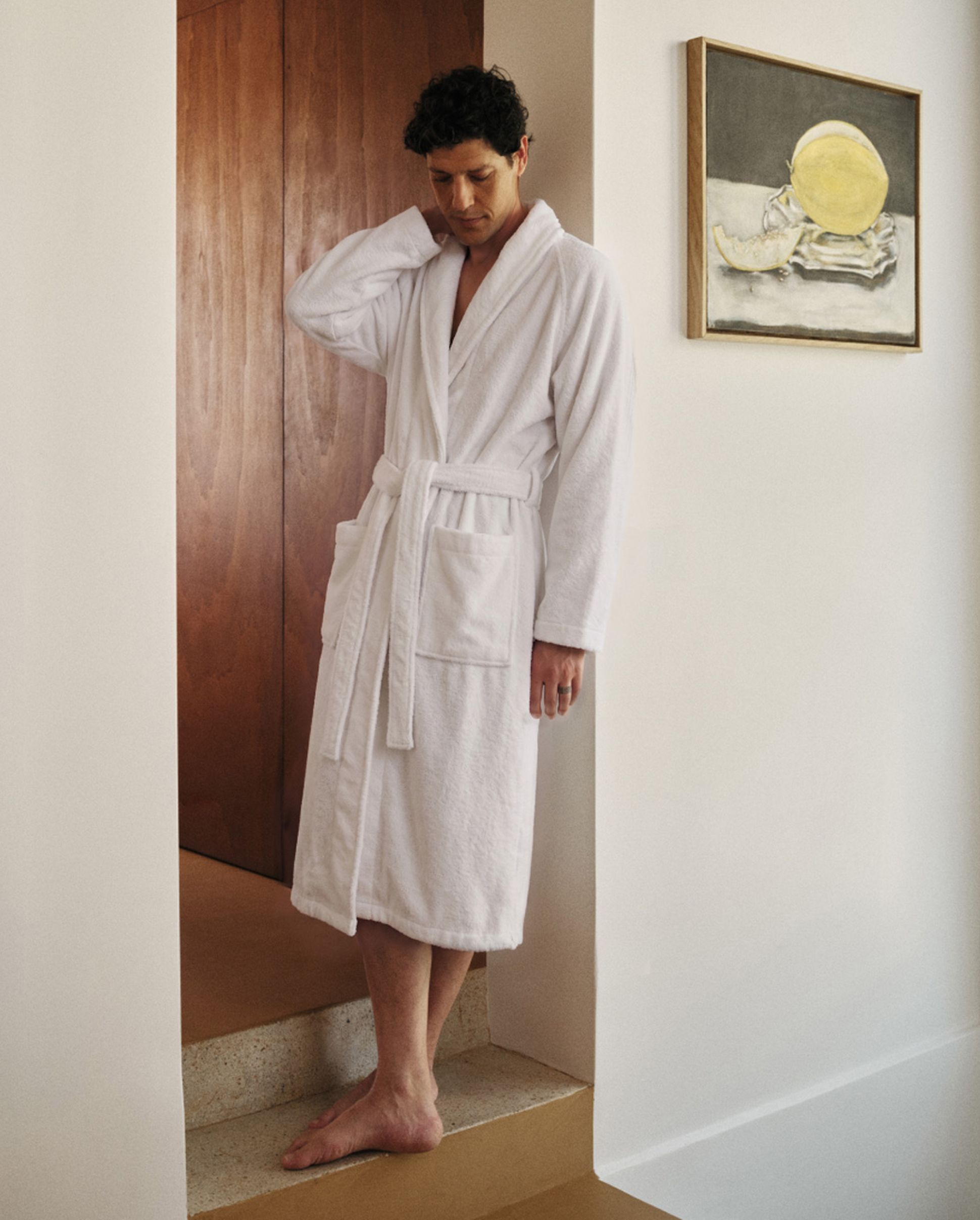 Man in a white bathrobe standing on steps in a modern interior with wooden door and lemon artwork on the wall.