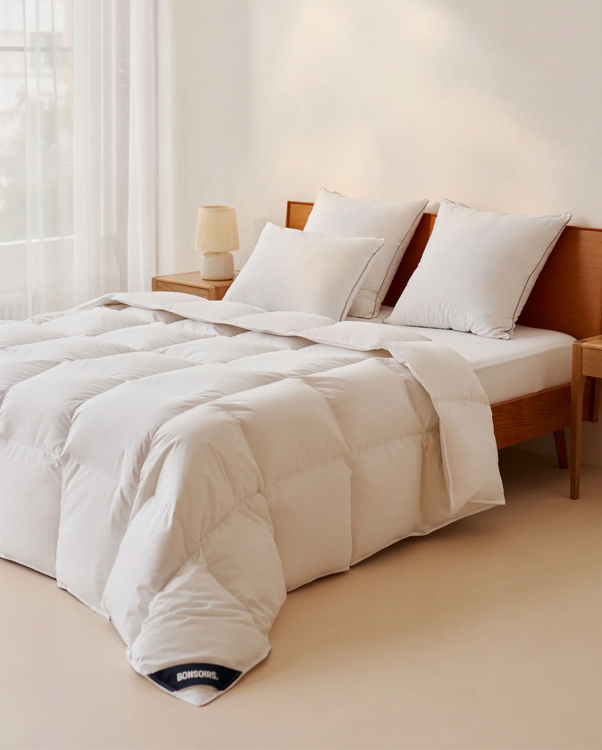 A neatly made bed with a fluffy white comforter, two pillows, and a wooden headboard, adjacent to a wooden nightstand with a lamp.