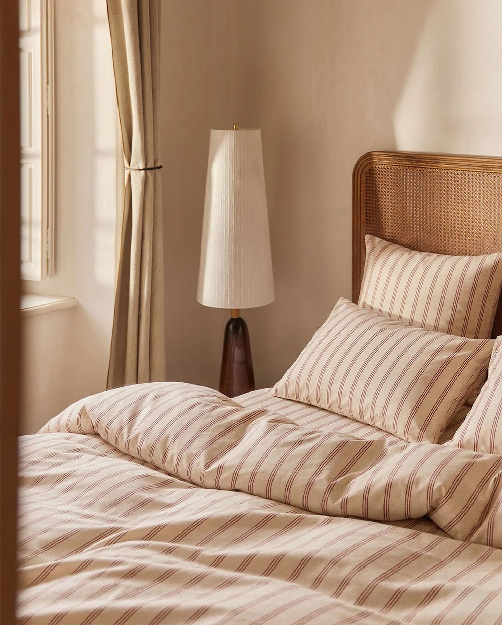 Une chambre douillette avec une parure de lit rayée, une tête de lit en rotin, un élégant lampadaire et des rideaux légers devant une fenêtre baignée de soleil.