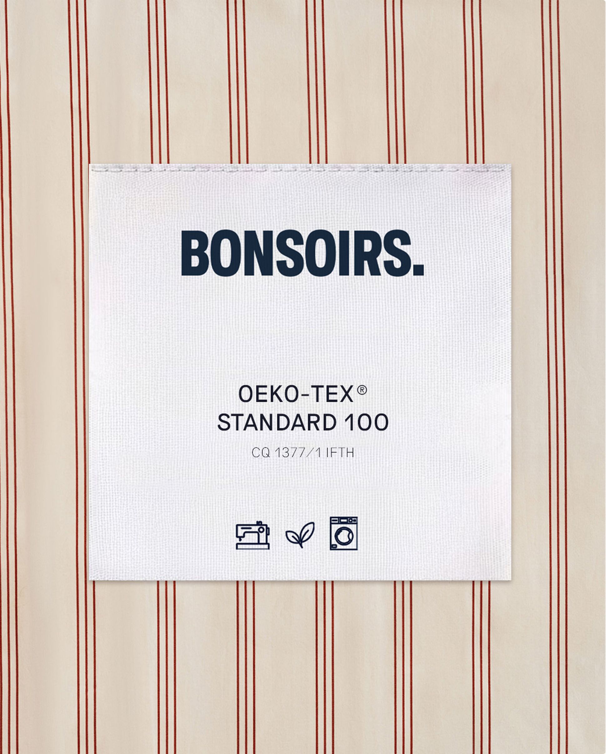 Une étiquette portant la mention « BONSOIRS. OEKO-TEX STANDARD 100 » est cousue sur un tissu à rayures verticales rouges. Des icônes indiquent les consignes d'entretien et les normes écologiques.