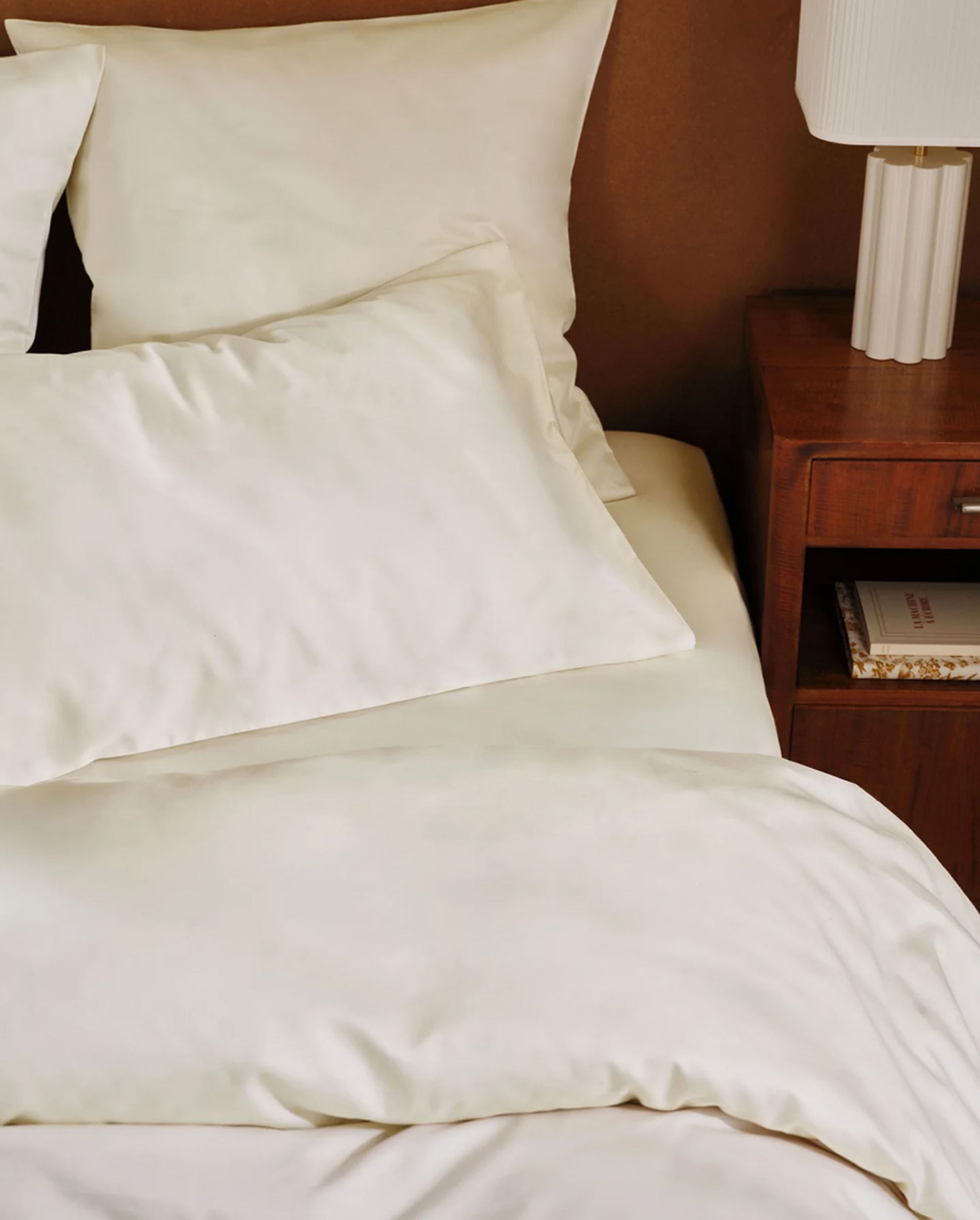 A neatly made bed with cream-colored sheets and pillows beside a wooden nightstand with a white lamp and a book.