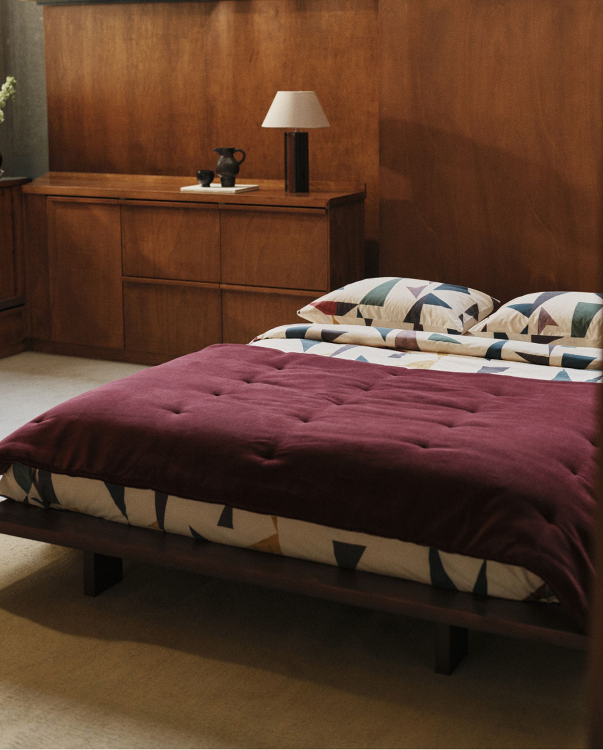 A cozy bedroom with a modern wooden bed featuring a geometric-patterned comforter and pillows. A dresser with a lamp and decor is in the background.
