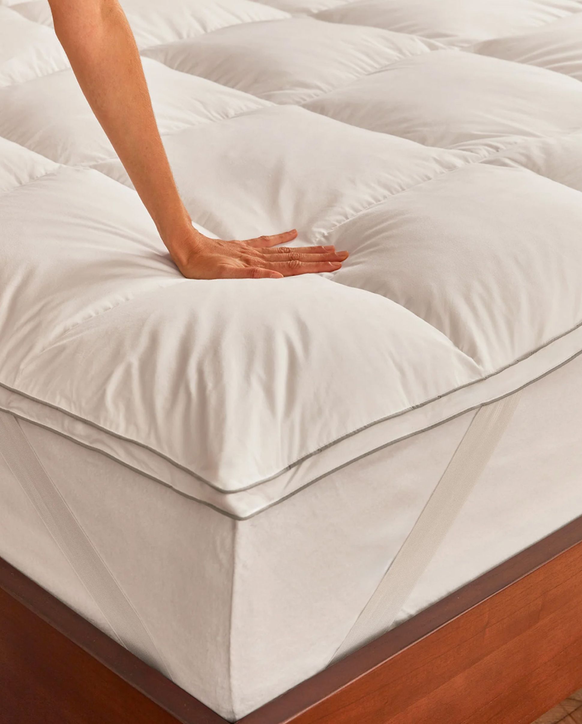 A hand gently pressing on a plush, white quilted mattress topper fitted on a wooden bed.
