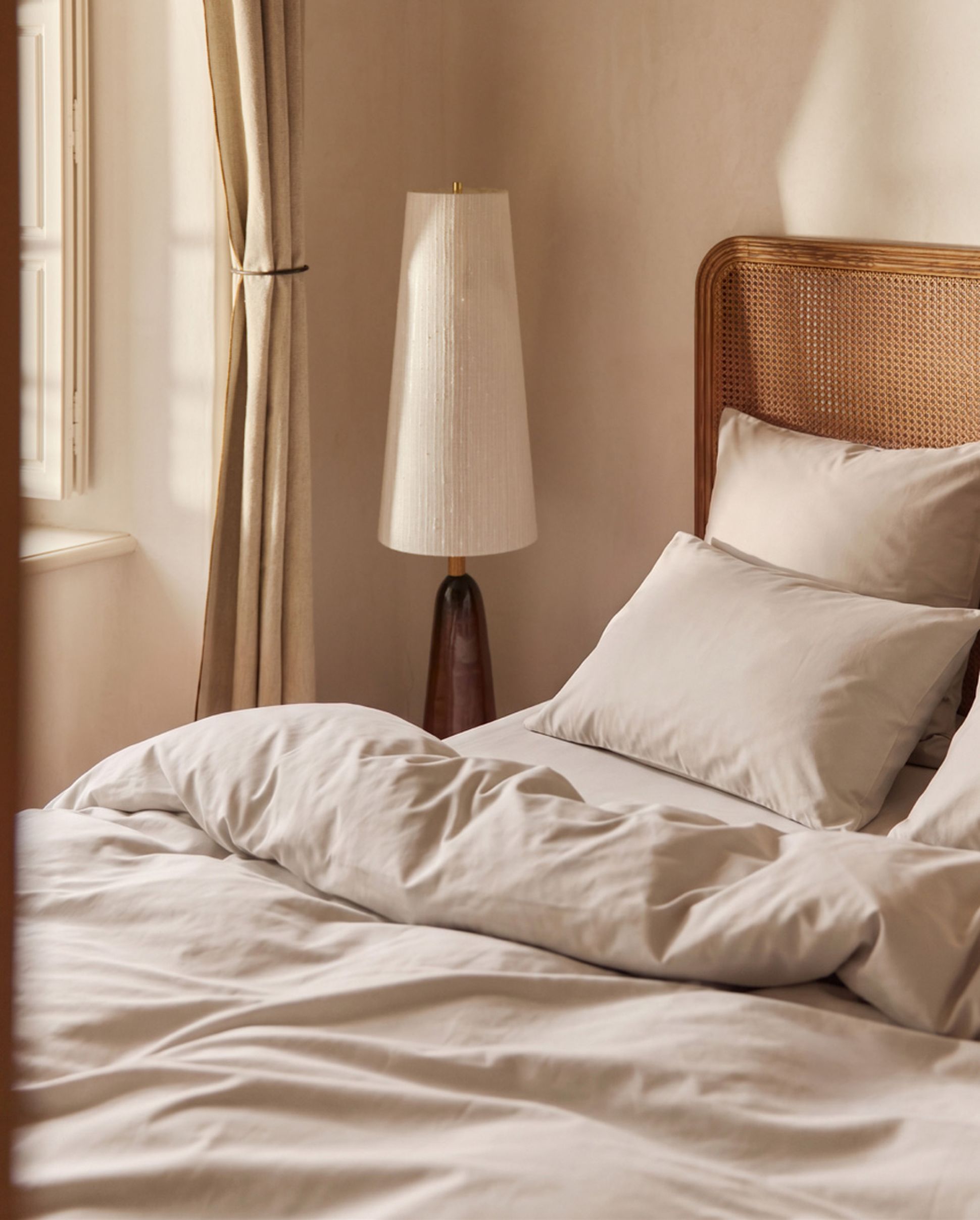 Cozy bedroom with a neatly made bed, white bedding, a modern lamp, and soft natural light filtering through curtains.