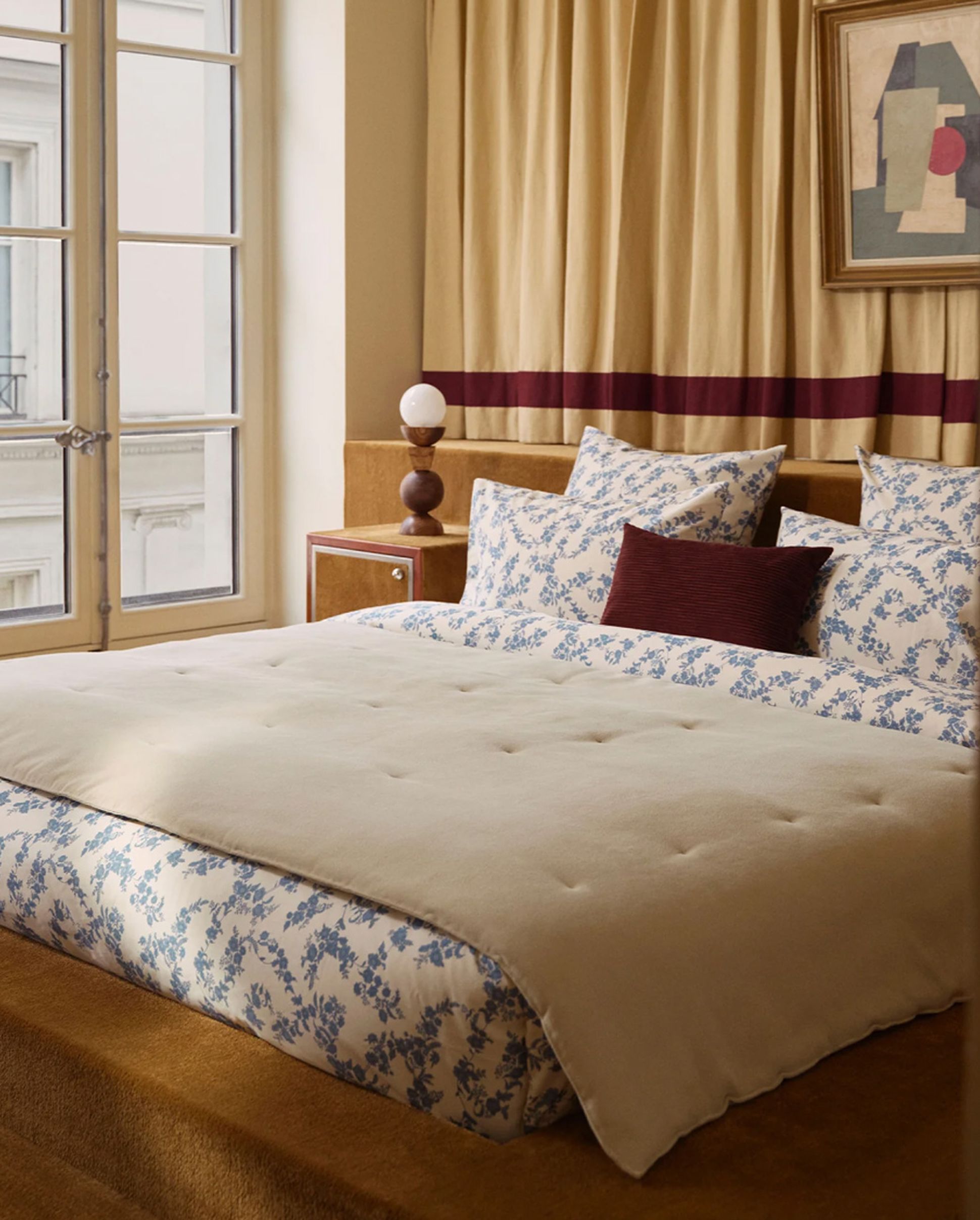 Elegant bedroom with a large bed, floral-patterned bedding, and a beige comforter. Drapes, artwork, and a nightstand with a lamp add warmth.