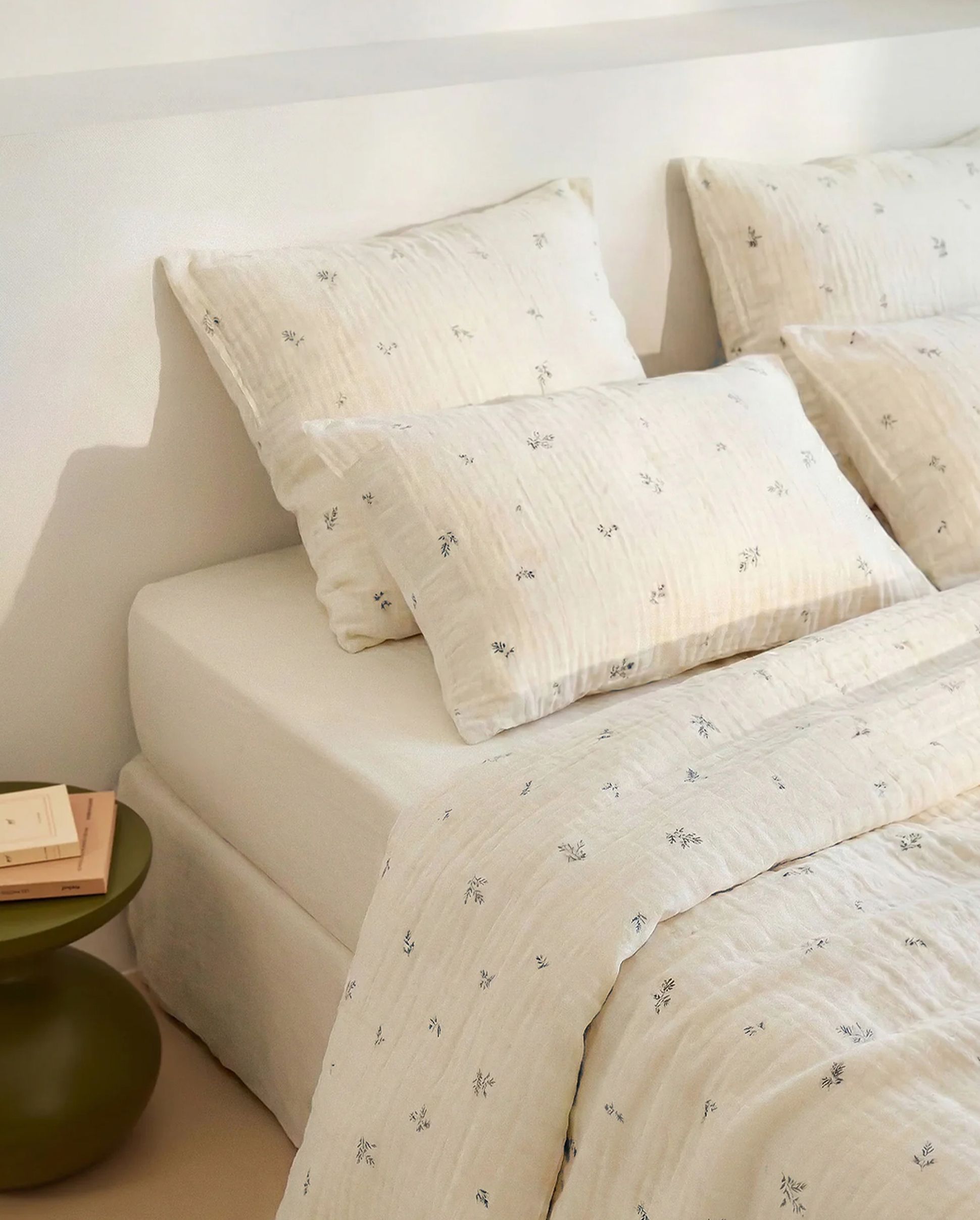 Un lit de couleur crème, avec des draps et des oreillers à motifs floraux, à côté d'une petite table de chevet verte sur laquelle sont posés des livres.