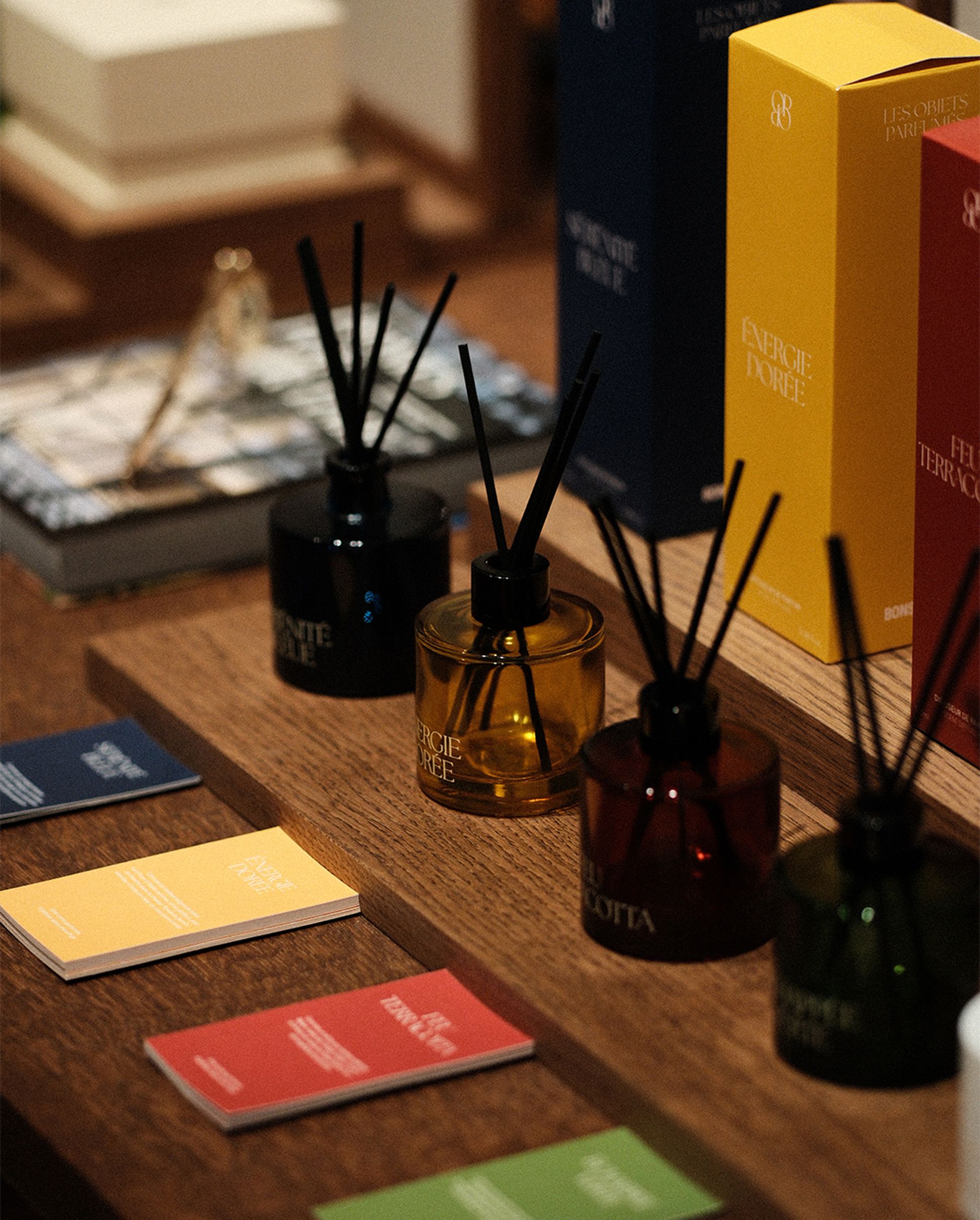 Une sélection de diffuseurs à bâtonnets de différentes couleurs et leurs emballages assortis, présentés sur un support en bois, avec des cartes de visite à proximité.