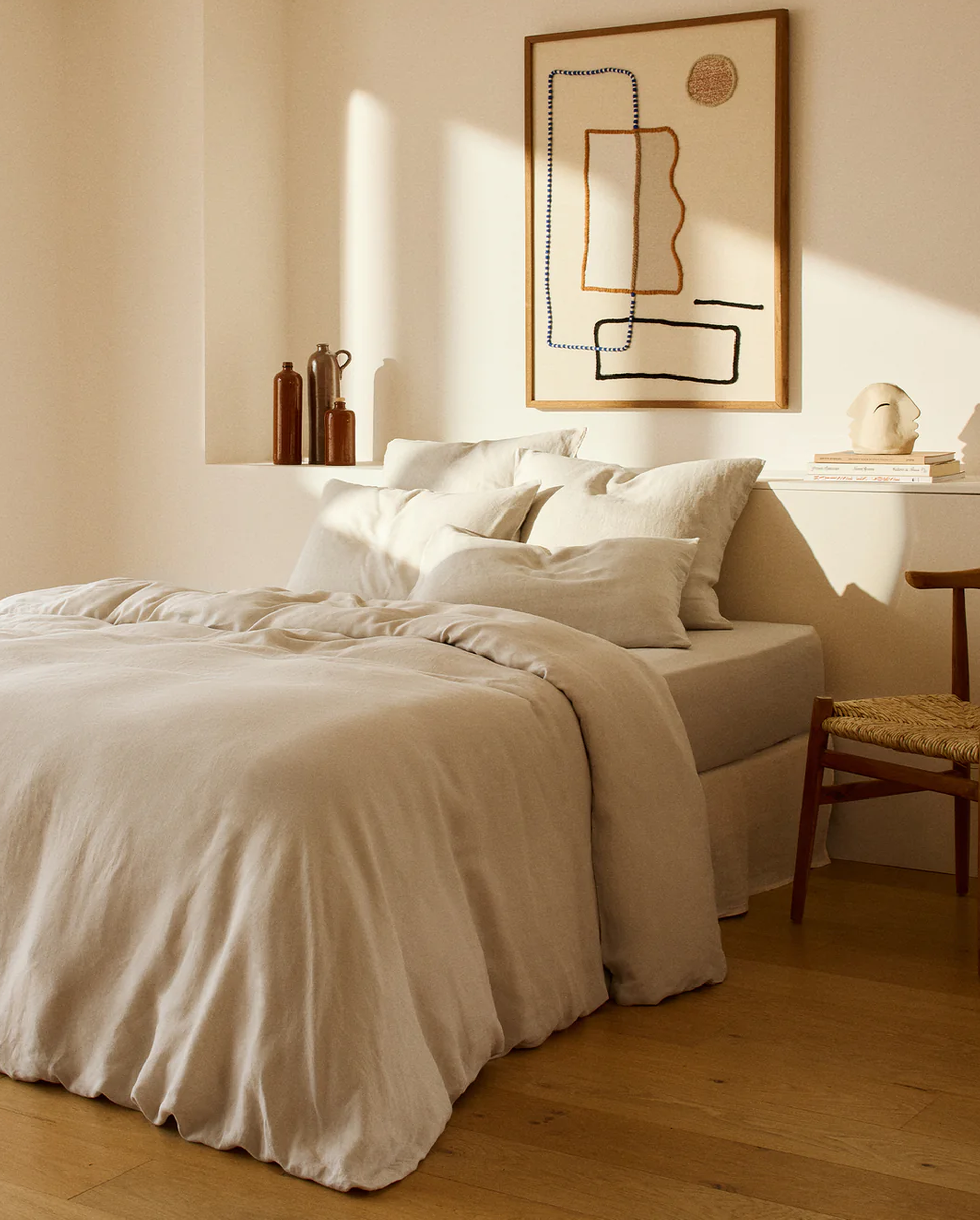 Une chambre minimaliste avec un lit beige, des oreillers blancs, des tableaux abstraits, des vases décoratifs, une petite sculpture et une chaise en bois posée sur un parquet.