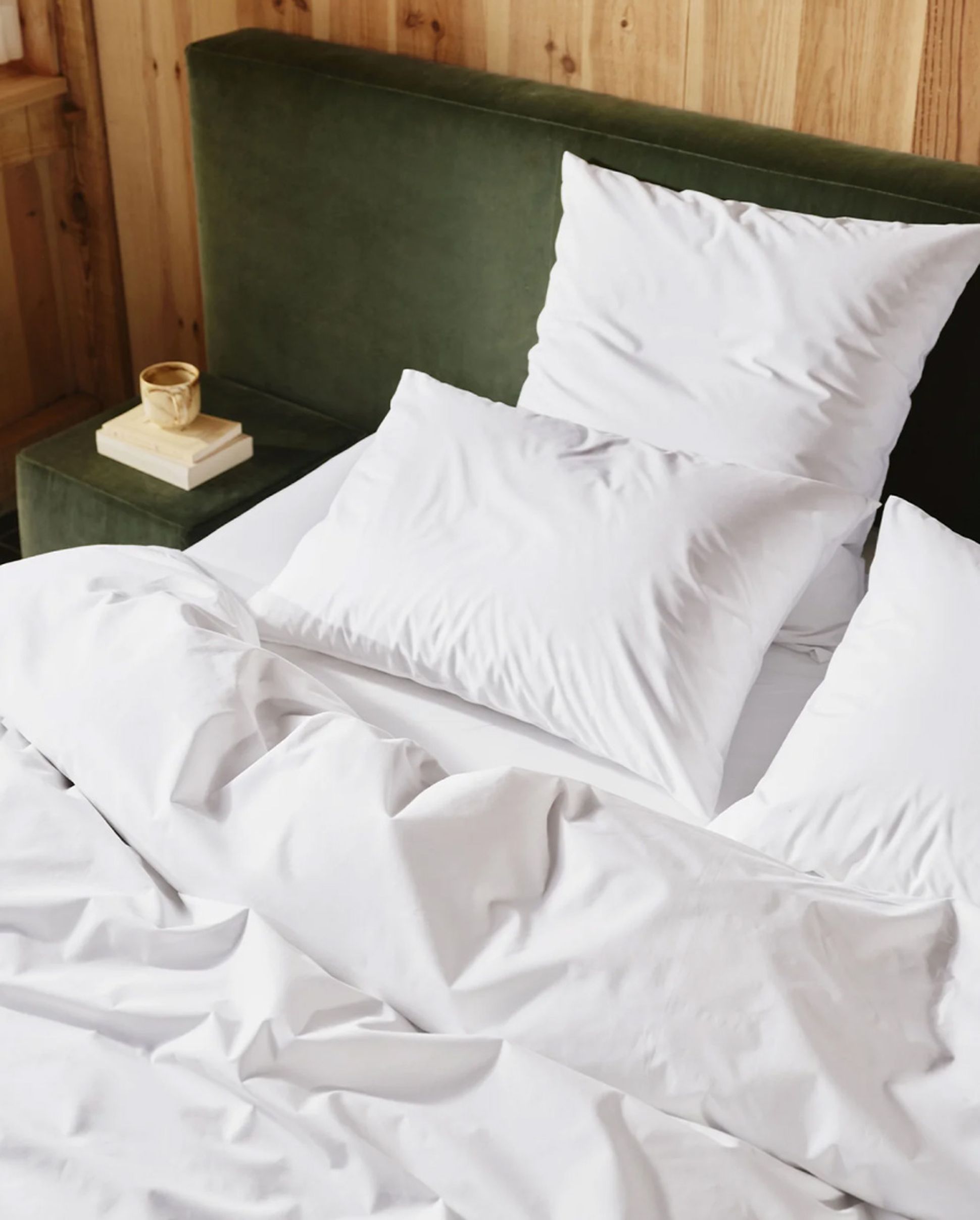 A neatly made bed with white pillows and sheets, a dark green upholstered headboard, and a wooden wall background; a mug on a book by the bed.