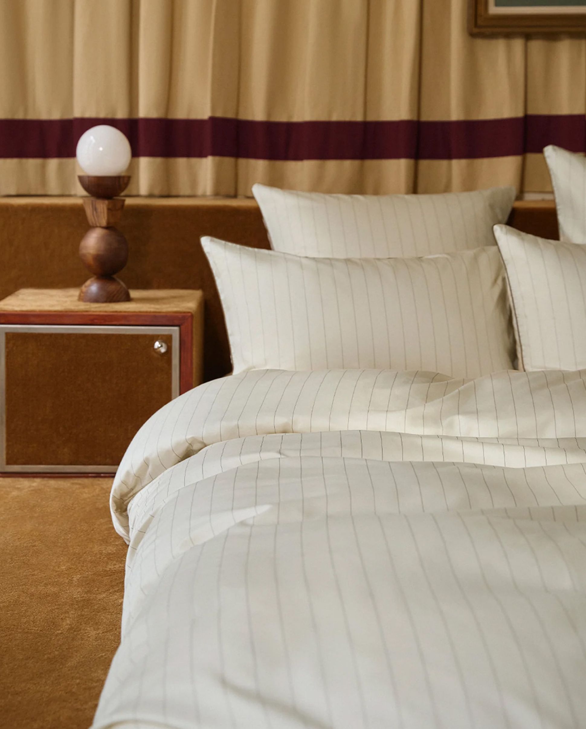 Bedroom with striped white bedding, several pillows, a wooden nightstand, a decorative lamp, and beige curtains with a maroon stripe.